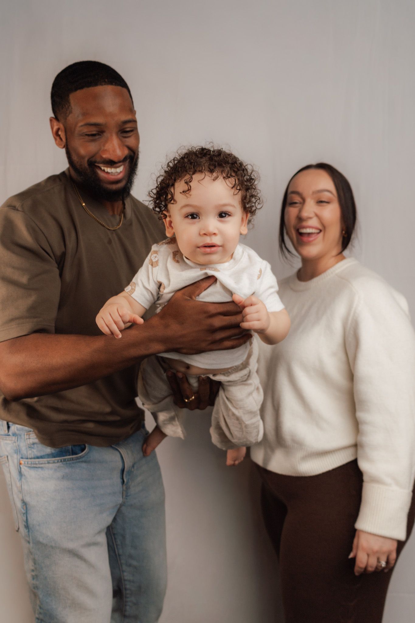 Happy family portrait with father holding curly-haired baby while mother laughs, against white background.