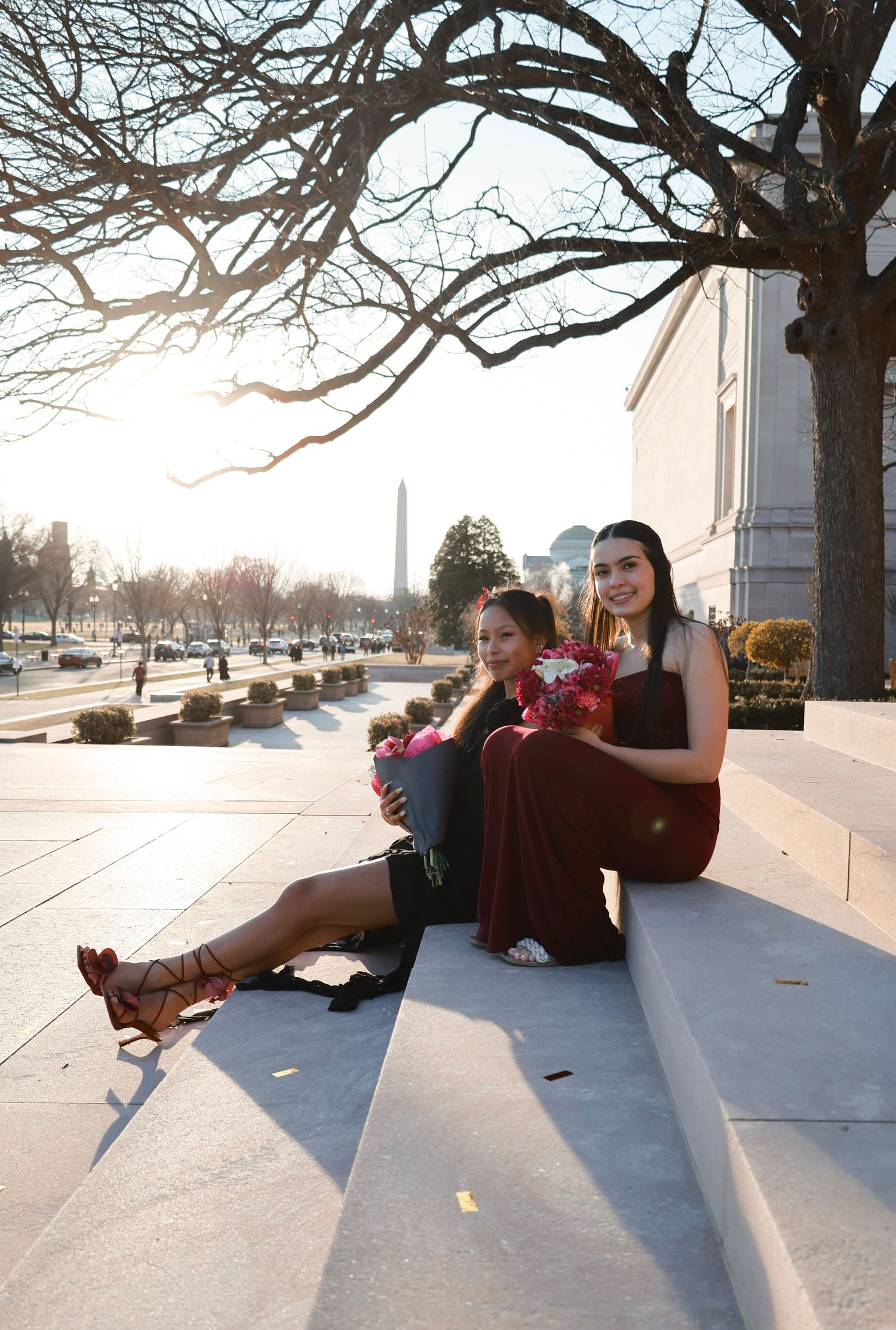 Two women in elegant burgundy dresses sit on steps near the Washington Monument on a sunny winter day.