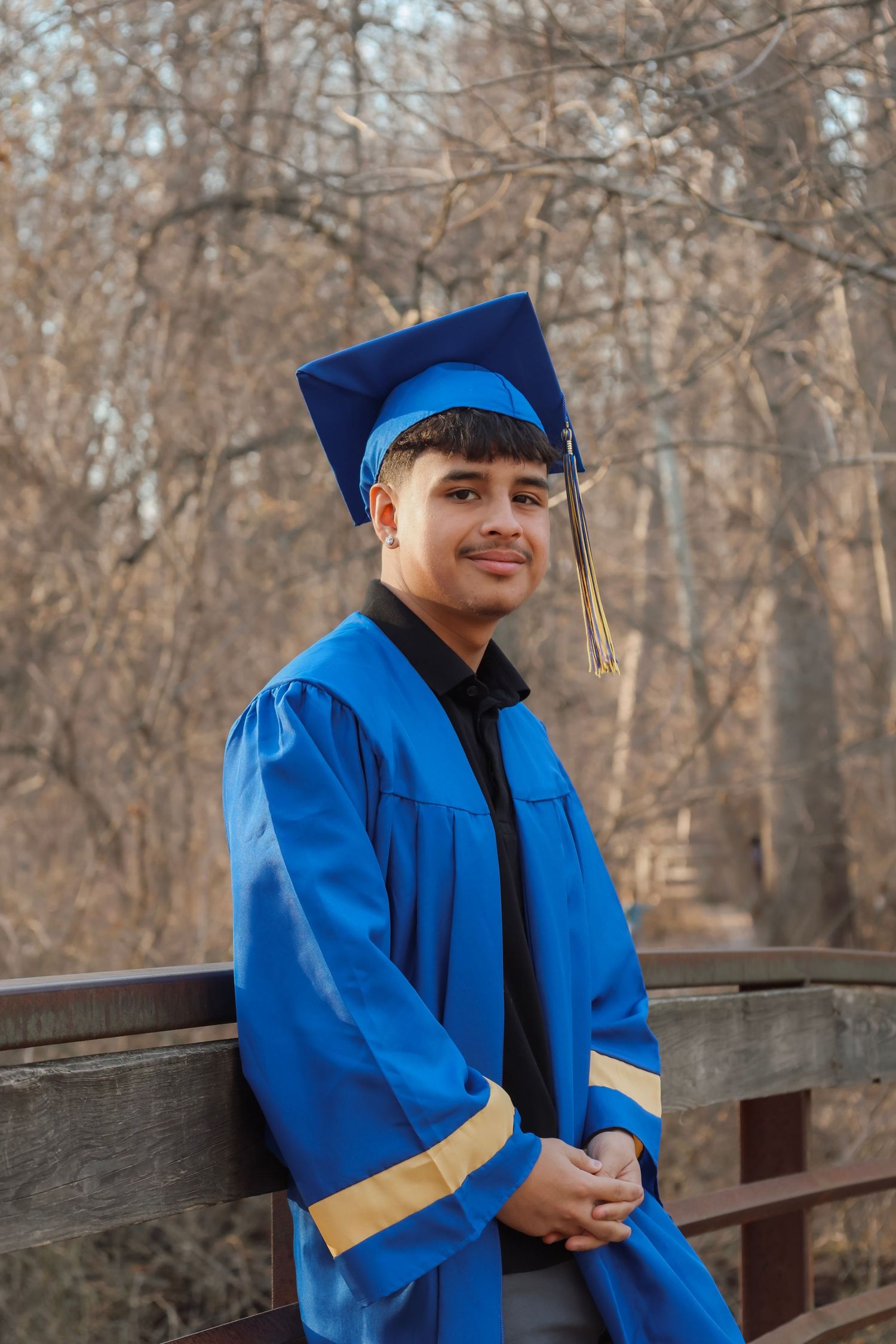 Young man in blue graduation cap and gown smiling on a wooden bridge surrounded by autumn trees.