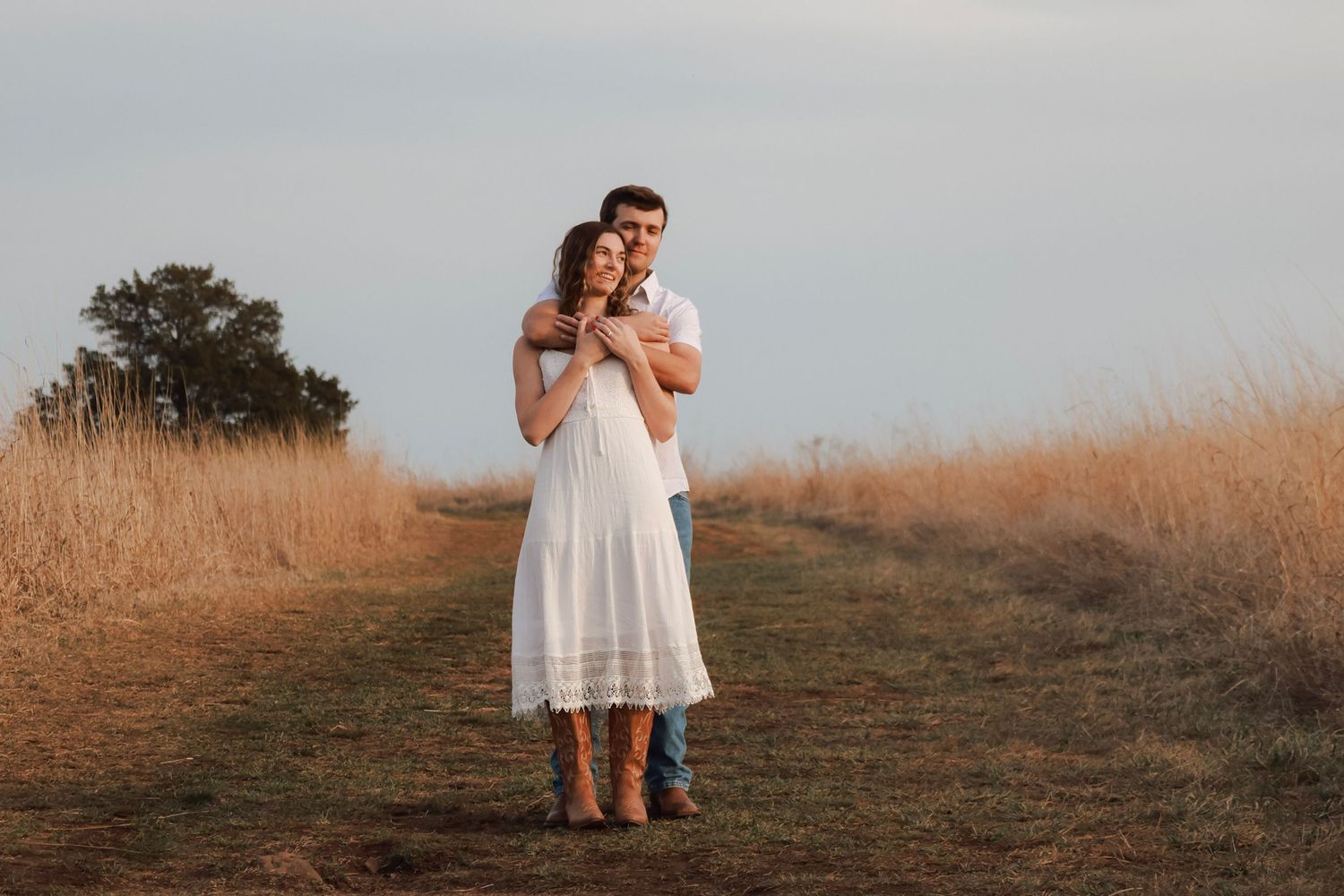 Couple embracing on a scenic dirt path surrounded by golden fields at sunset, woman in white dress.