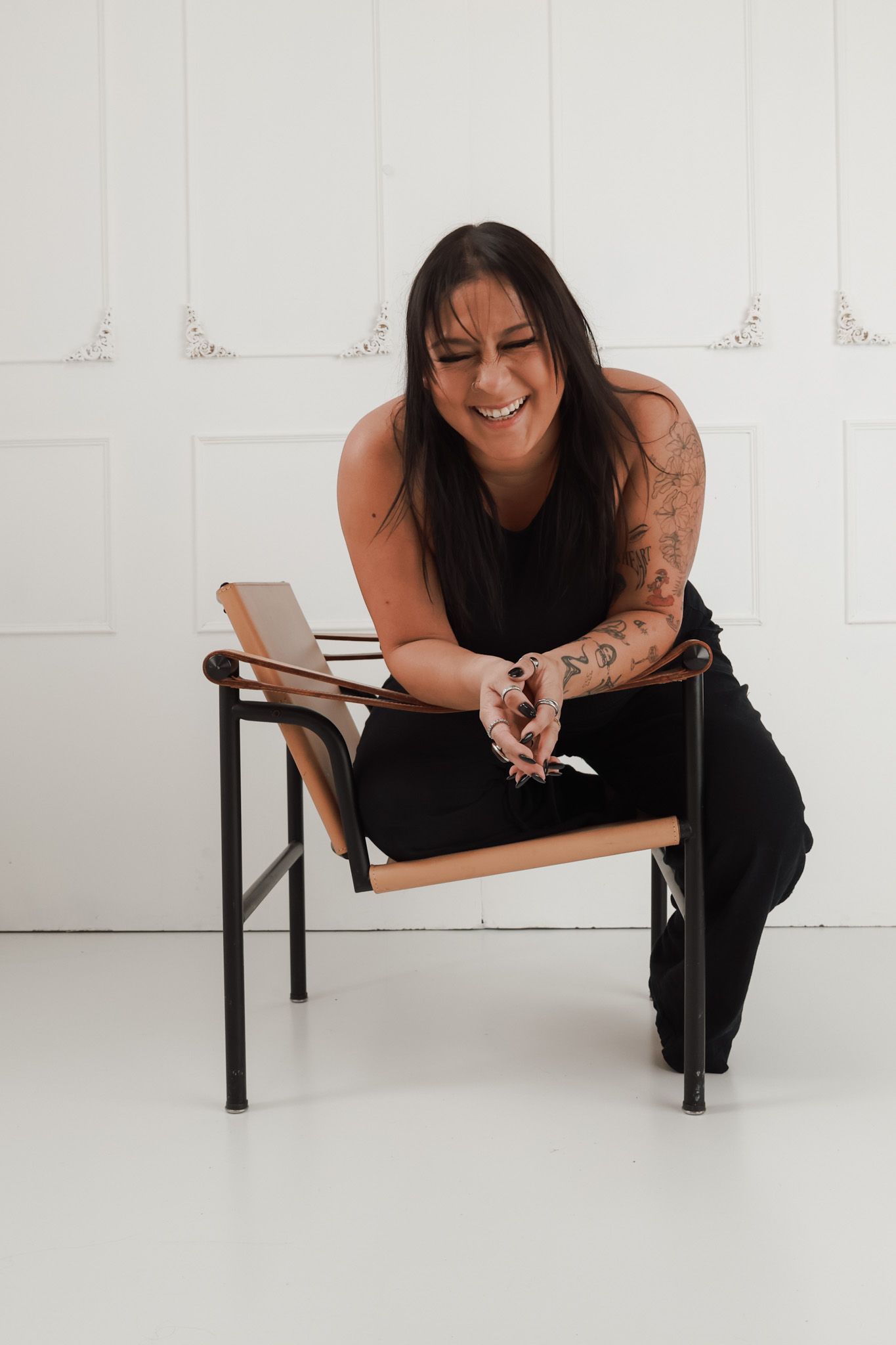 Tattooed woman in black outfit sitting on a modern chair, smiling and laughing in a bright white studio.