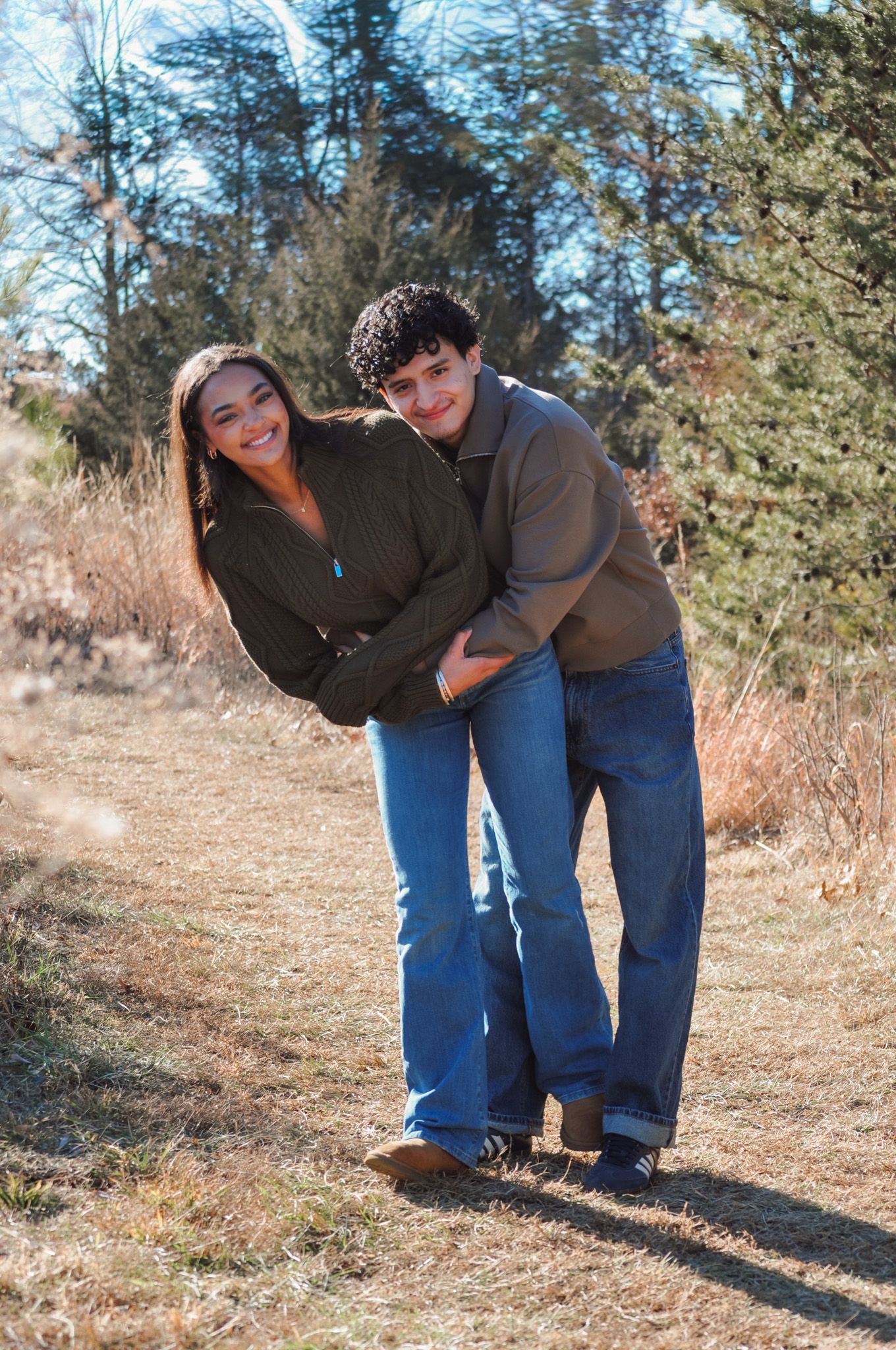 A young couple smiling and embracing outdoors on a sunny day, wearing matching brown sweaters and blue jeans.