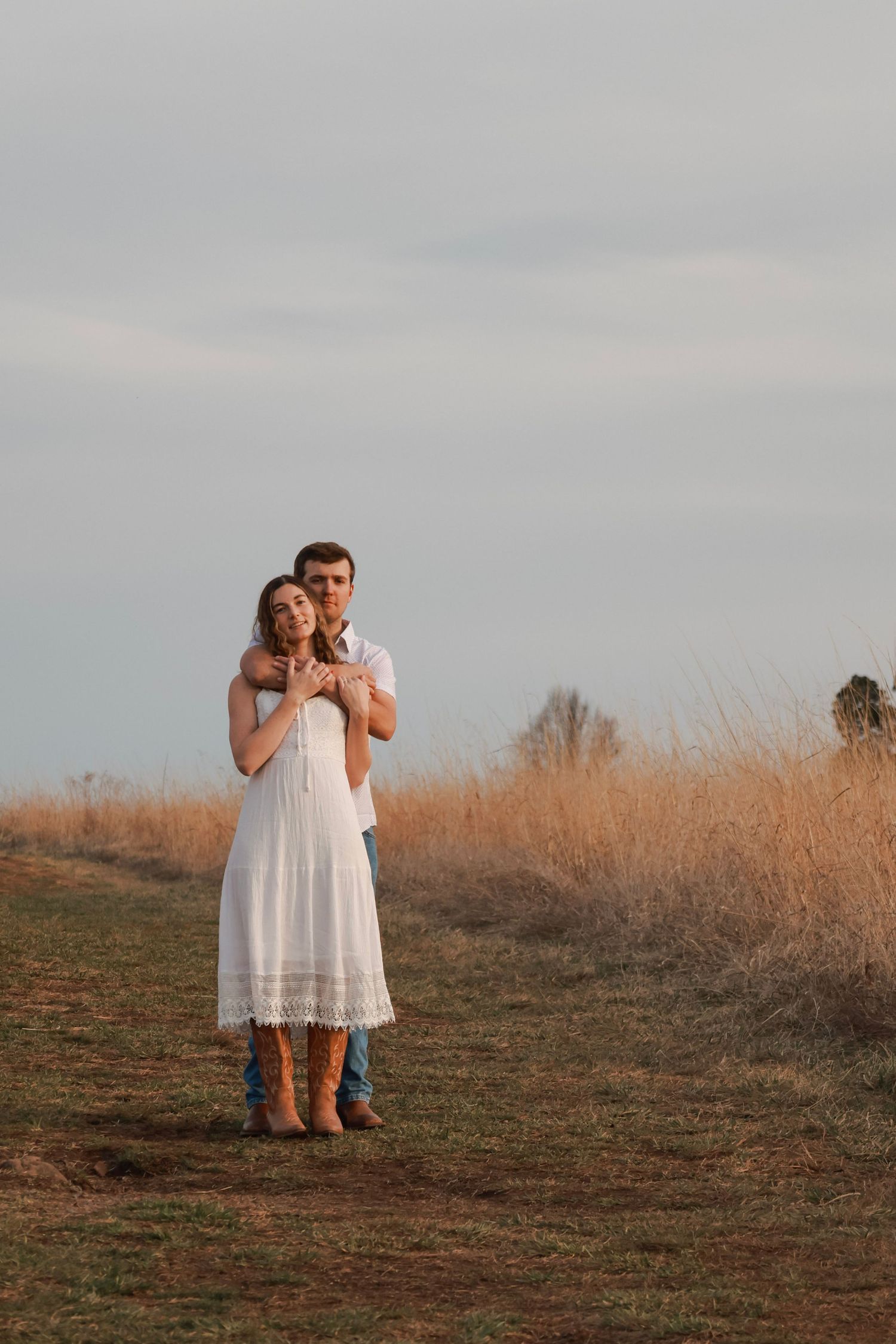 Couple embracing in a golden field, woman in white lace dress and boots, man in blue jeans, under a cloudy sky.