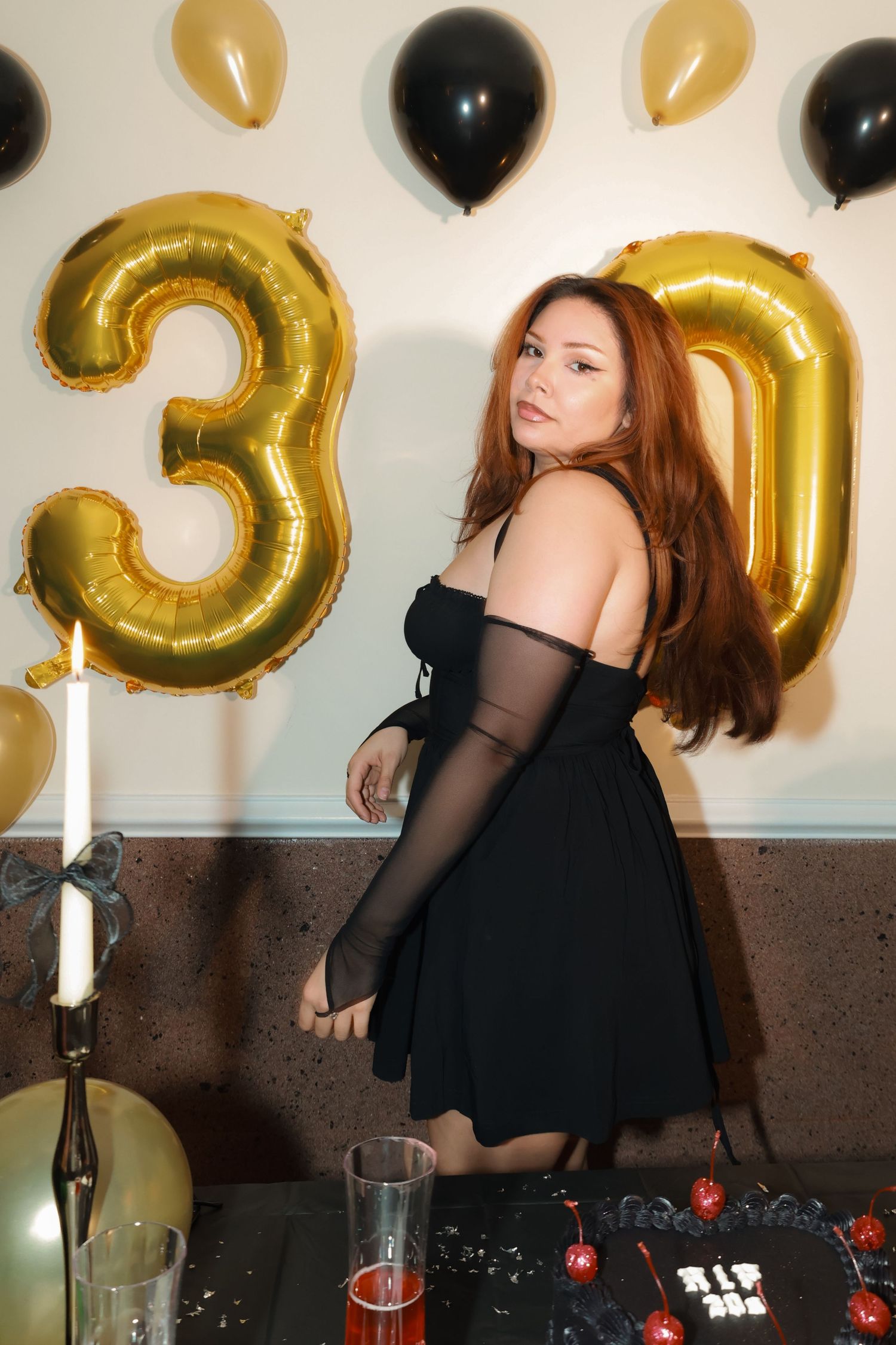 Woman in black off-shoulder dress poses at 30th birthday party with gold balloons and decorated table.