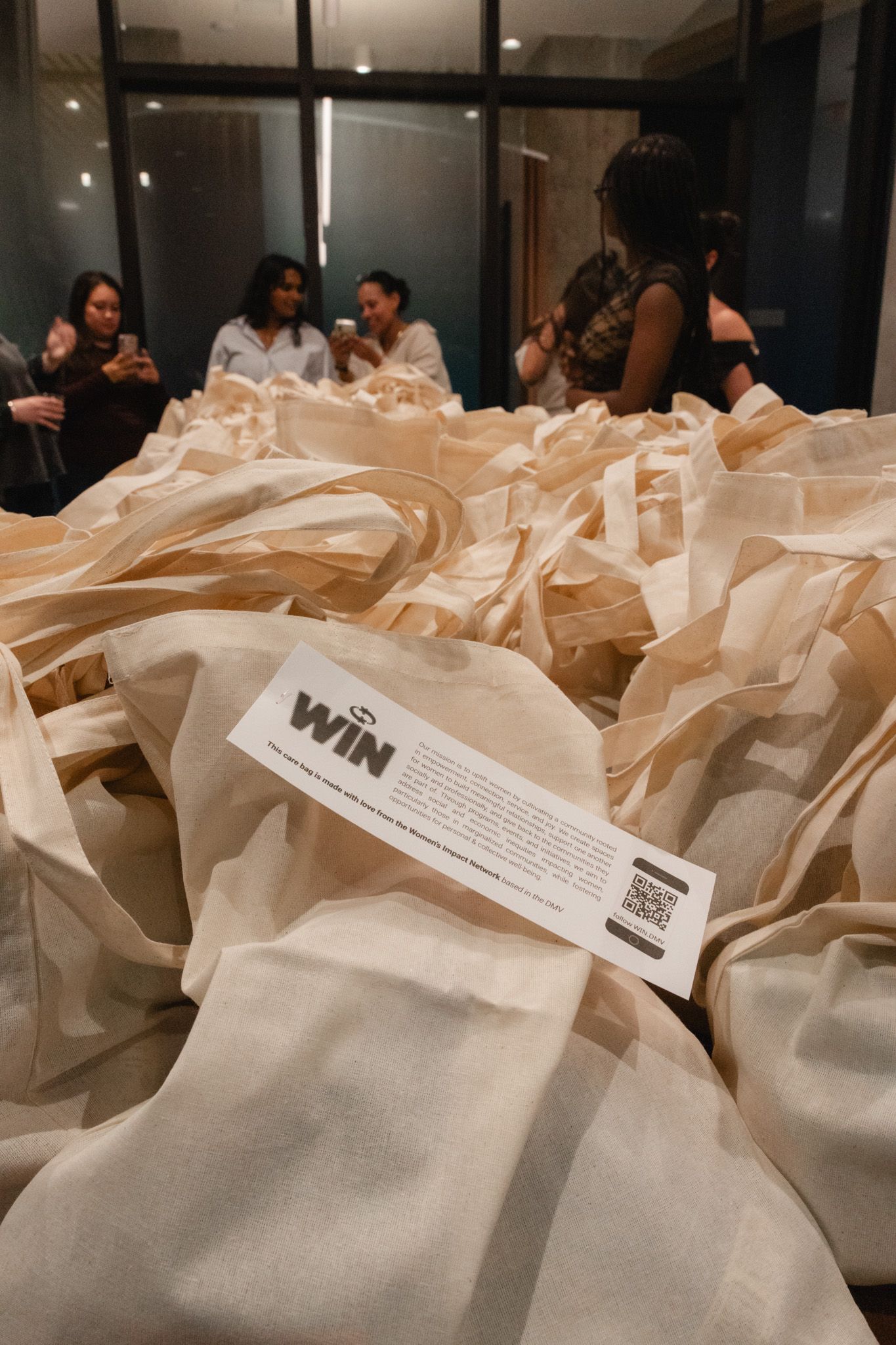Pile of cream canvas tote bags with a WIN promotional card at an indoor event with women in background.