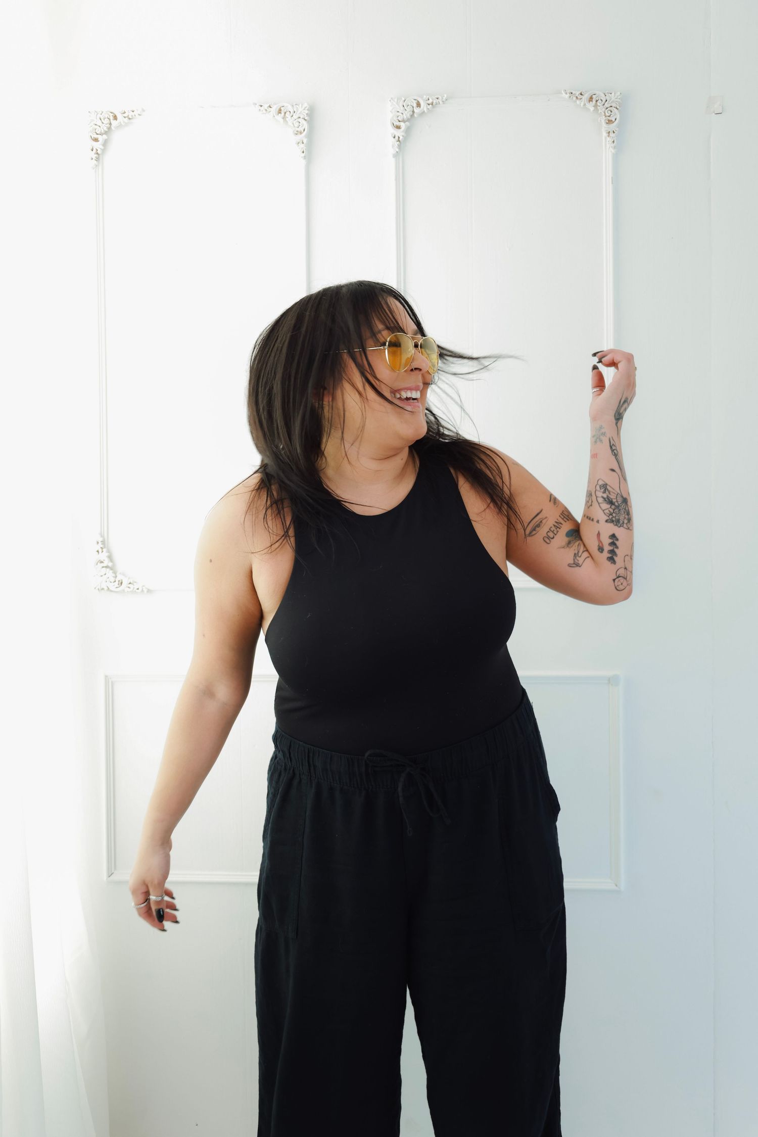 Smiling woman in black tank top and pants with tattoos and yellow sunglasses posing against white wall in a studio.
