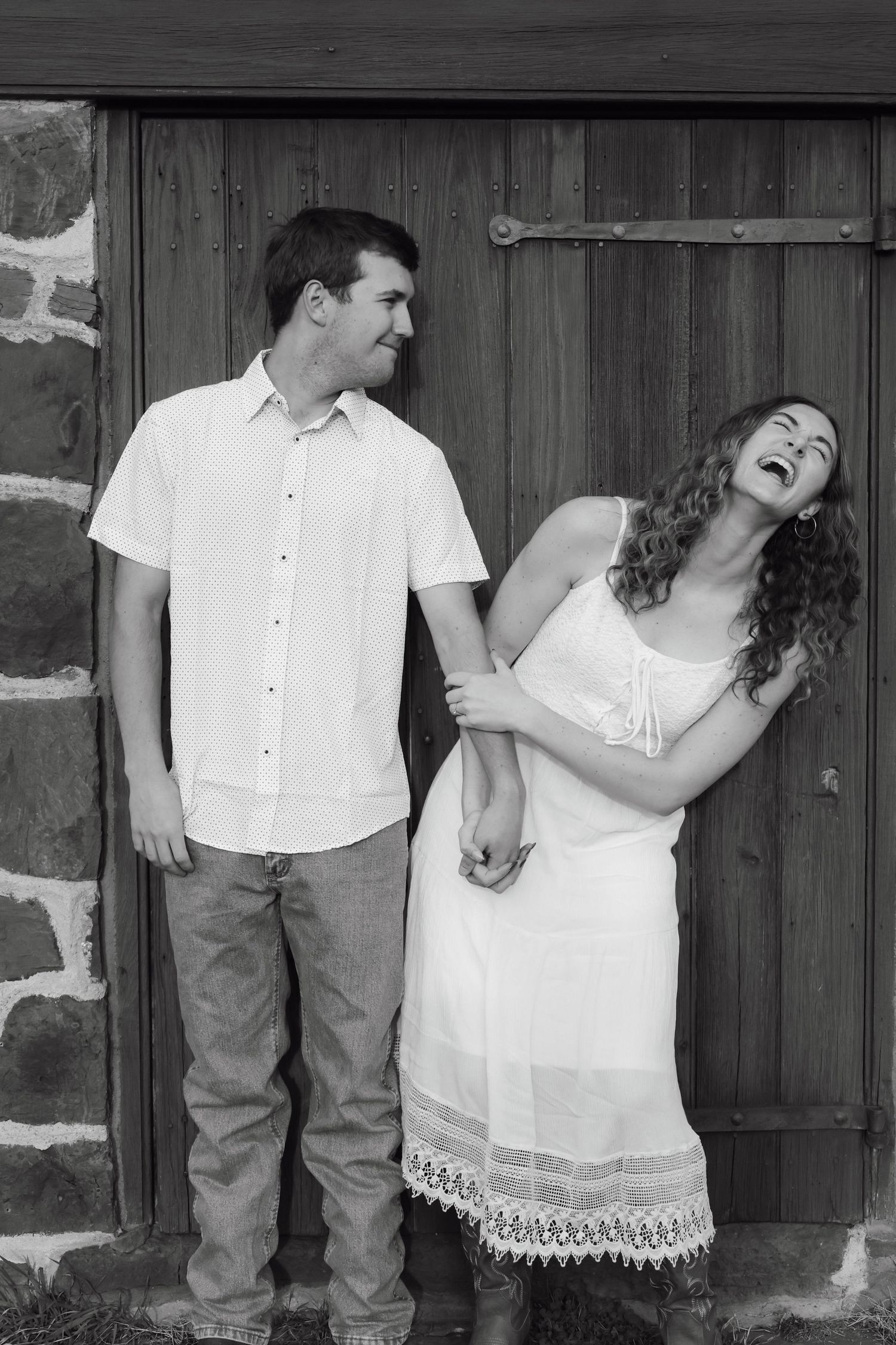 Black and white photo of a laughing couple holding hands in front of a rustic wooden door and stone wall.