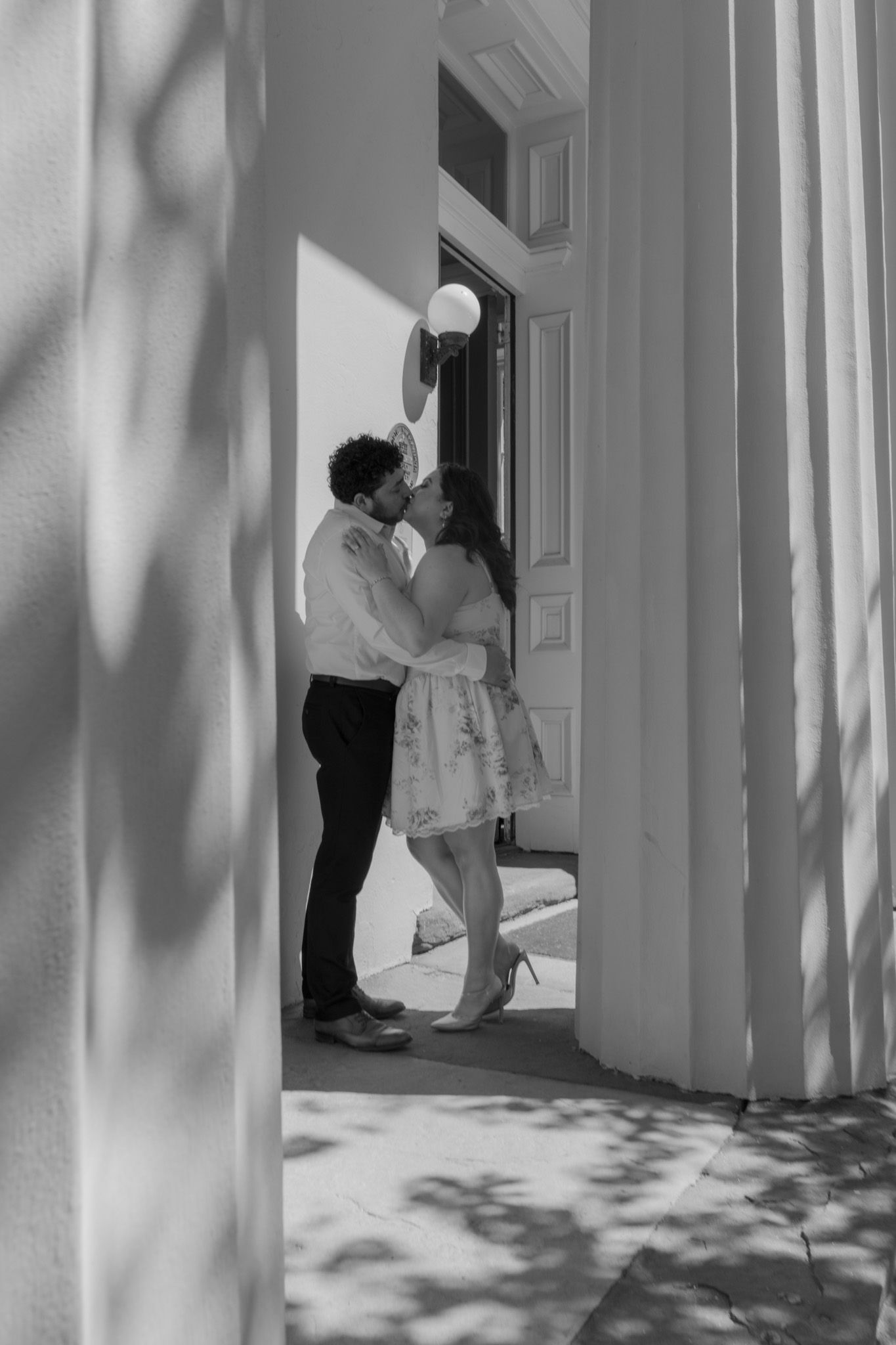 Black and white photo of a couple sharing a romantic kiss in a sunlit doorway between white columns.
