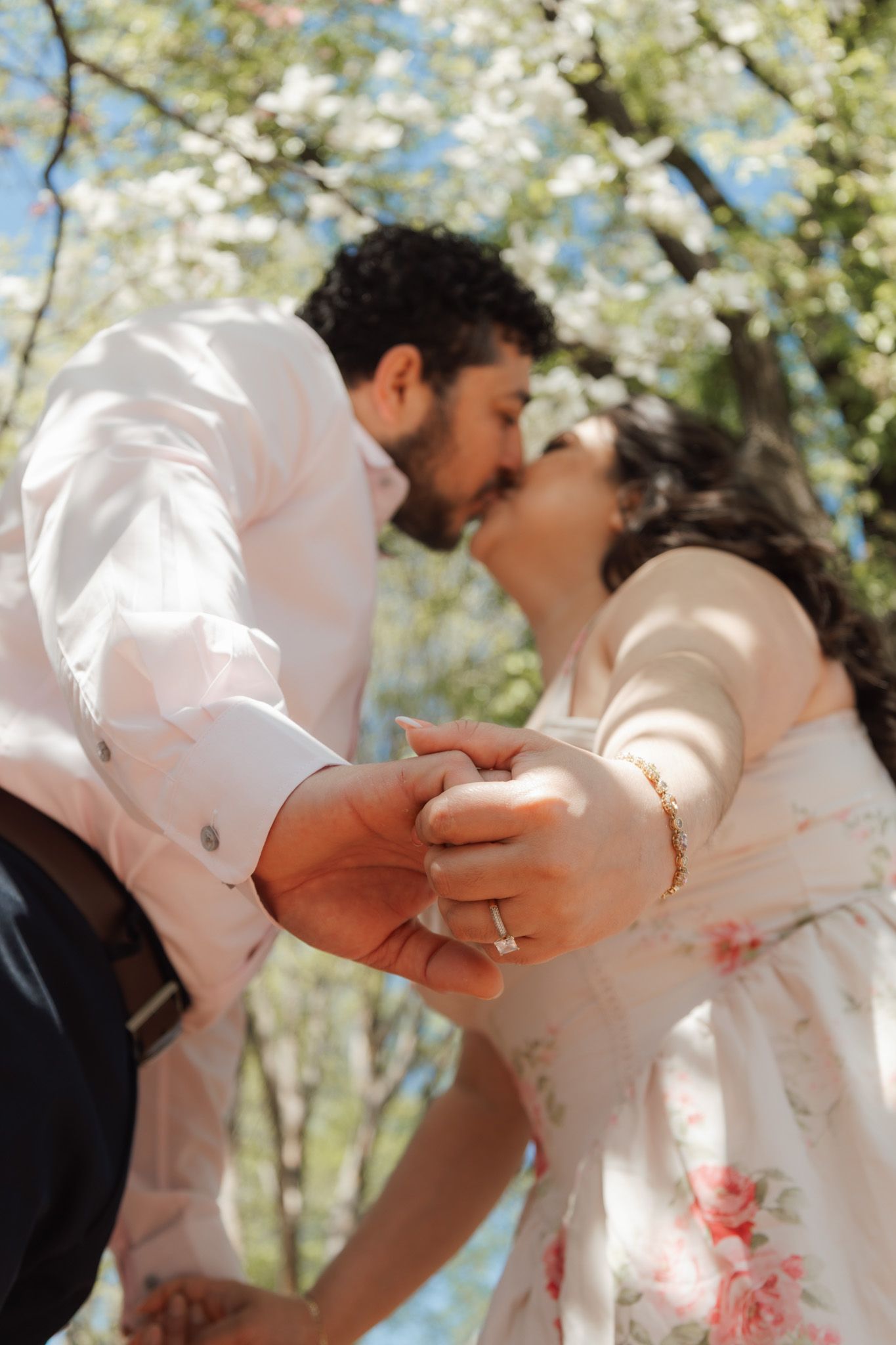 Couple sharing a romantic kiss under blooming white cherry blossom trees, holding hands with an engagement ring visible.