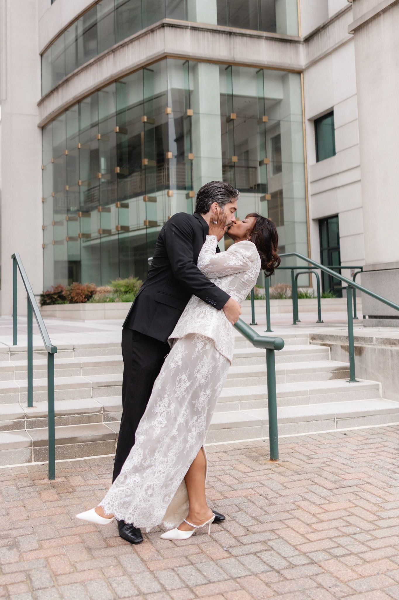 Couple sharing a romantic kiss on courthouse steps, bride in white lace gown and groom in black tuxedo.