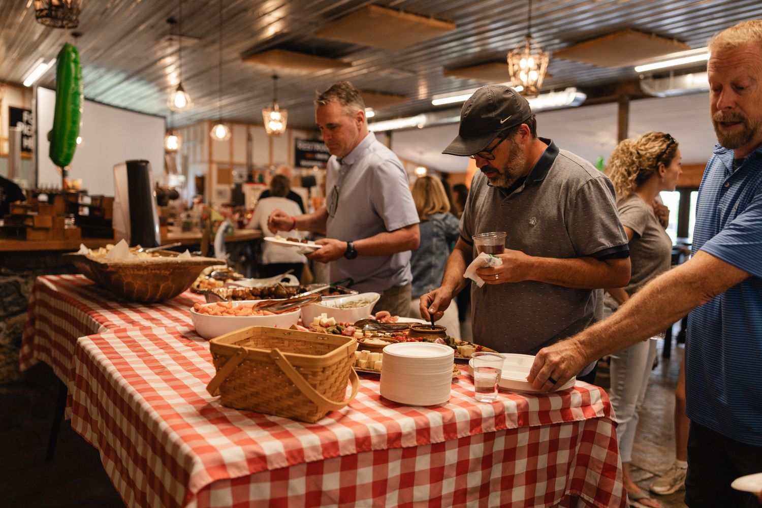 Event photography with People serving themselves at a buffet table with red checkered tablecloth at a casual indoor gathering.