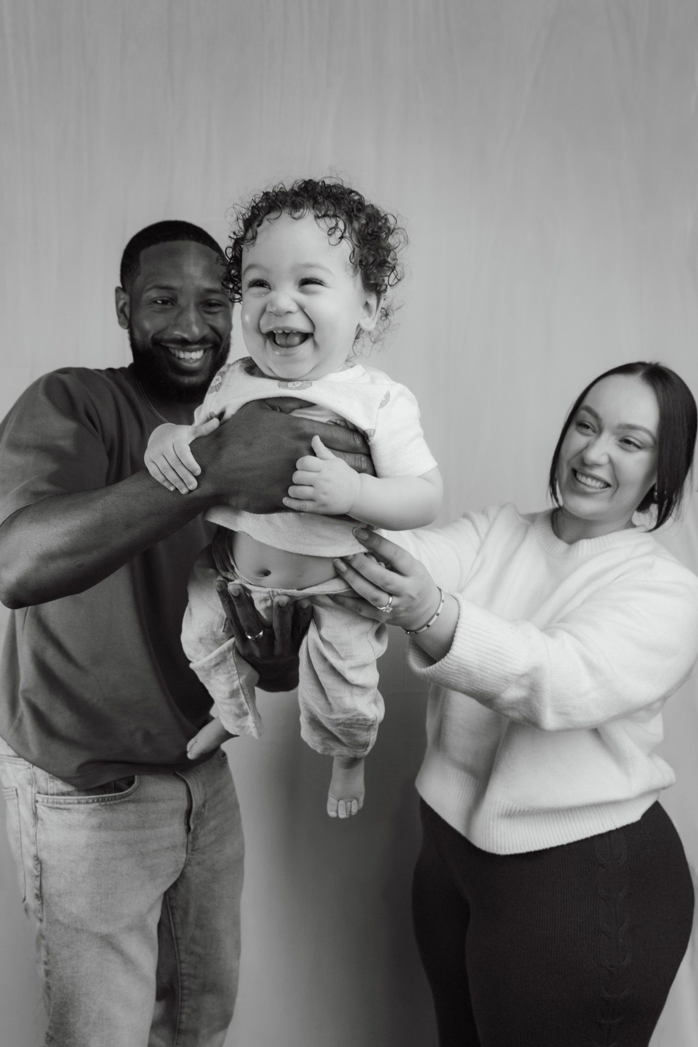 Black and white photo of smiling parents lifting their laughing baby together in a studio portrait.