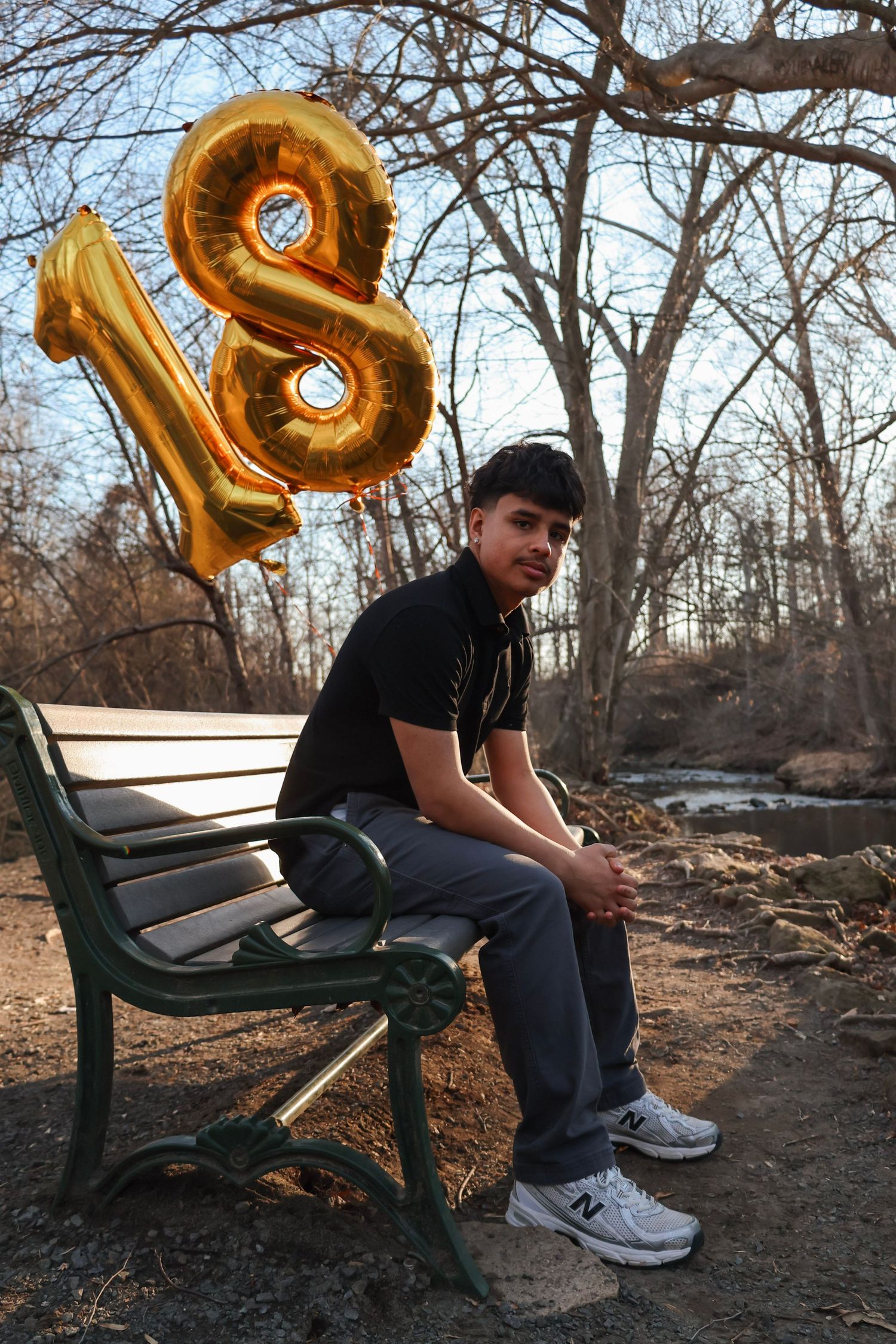 Young man sitting on a park bench celebrating his 18th birthday with gold number balloons outdoors.
