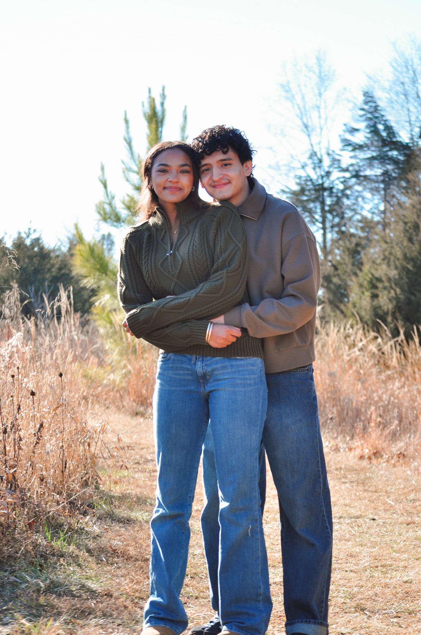A young couple embracing outdoors in fall, wearing earth-tone sweaters and jeans, surrounded by dry grass and trees.