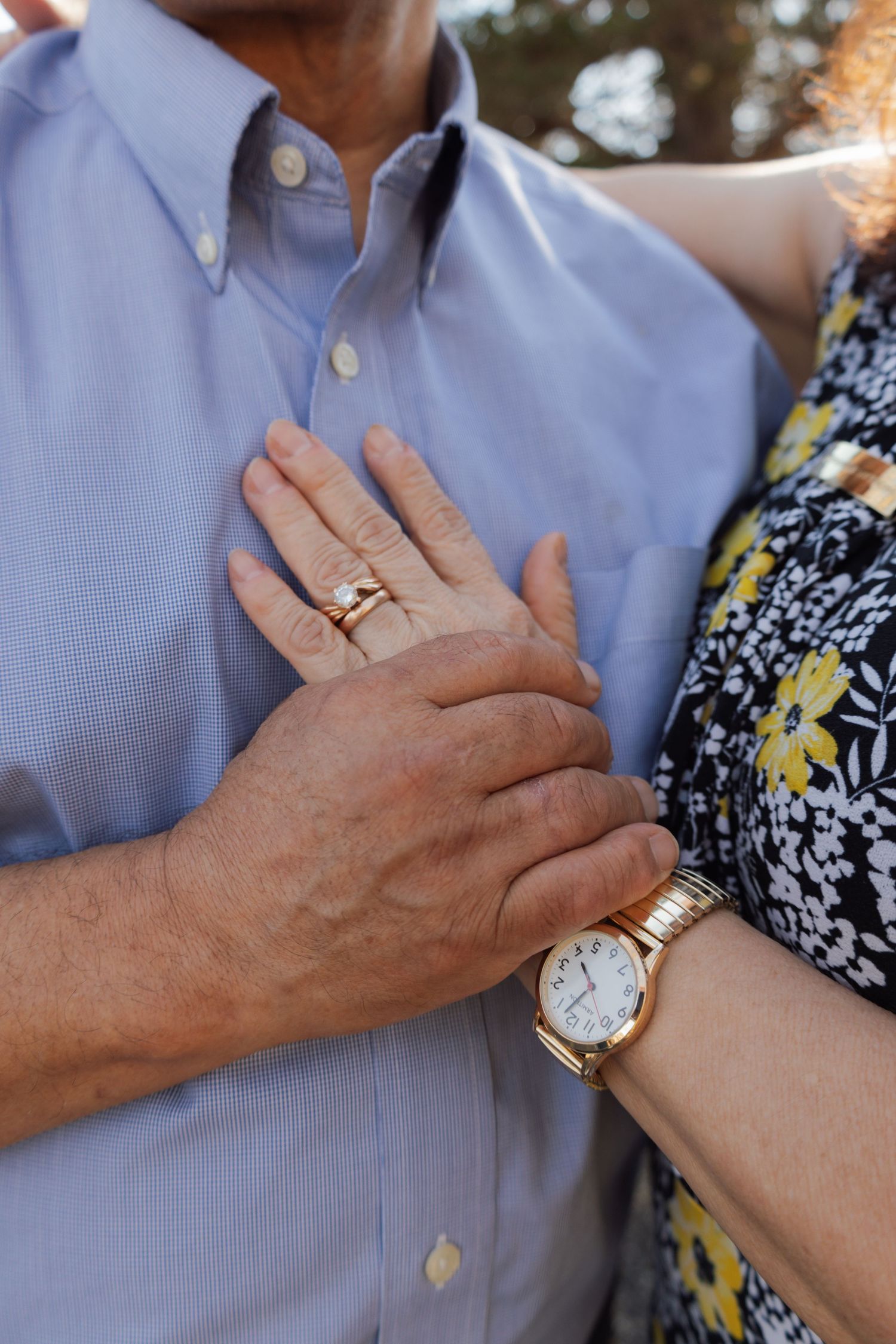 Close-up of an older couple's hands, woman wearing a gold watch and rings, resting on man's blue button-up shirt.