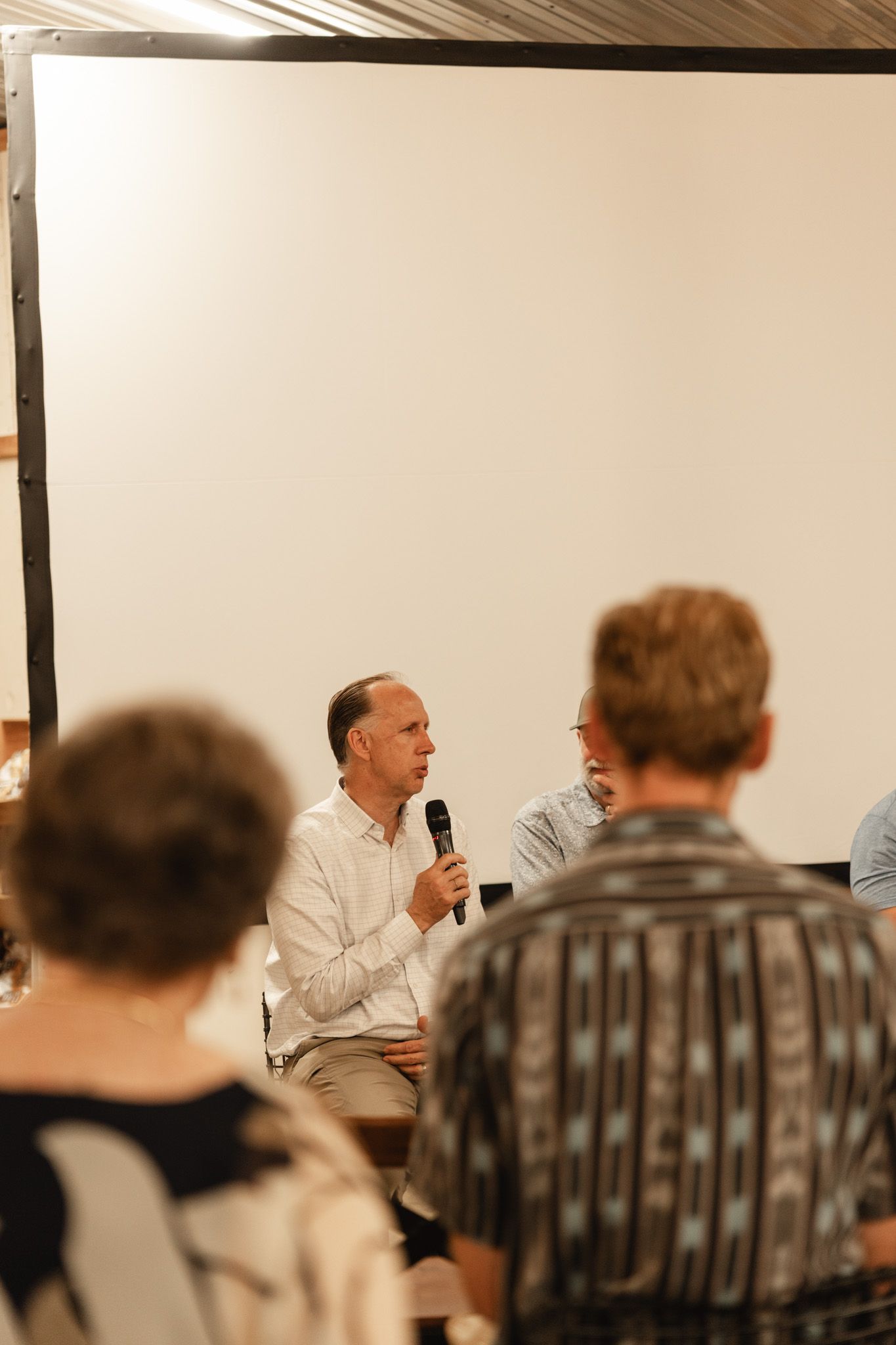 Man in white shirt speaks into microphone at indoor panel discussion with audience members in foreground.
