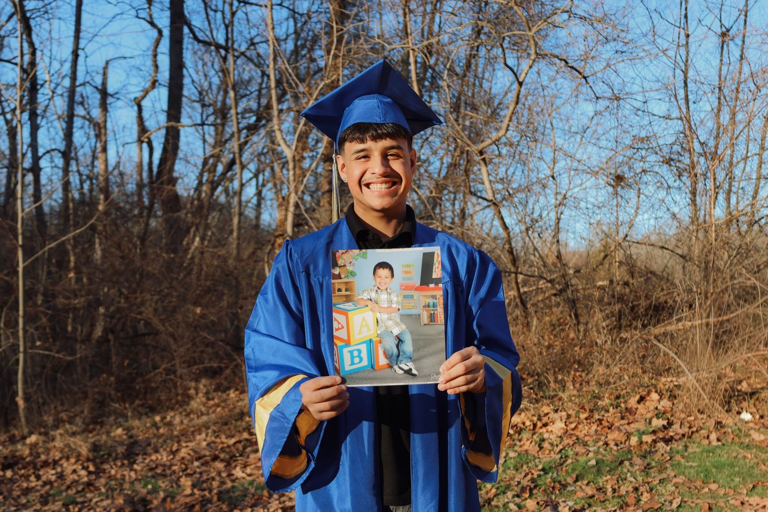 Smiling graduate in blue cap and gown holds childhood photo outdoors among autumn trees.