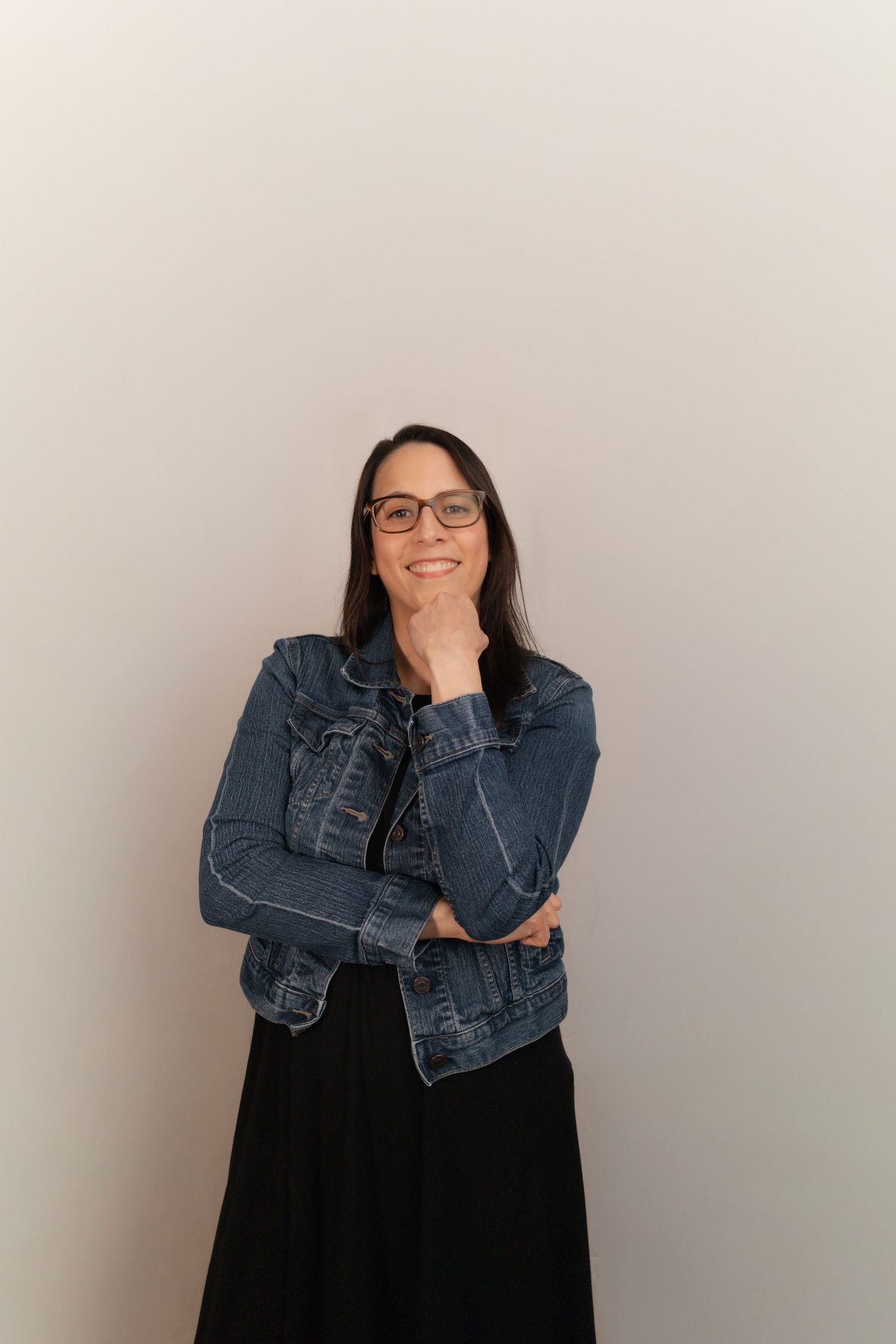 Smiling woman in denim jacket and glasses posing with hand on chin against a light gray background.