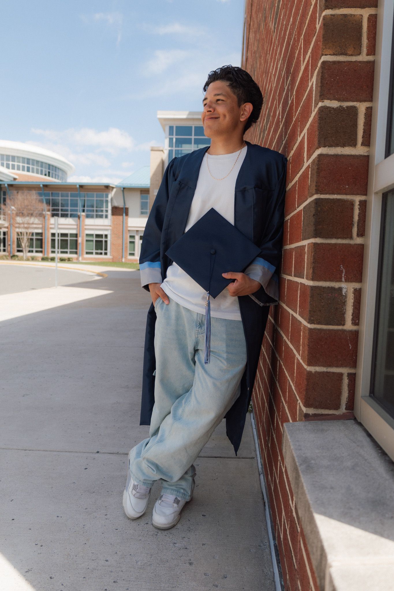 Young graduate in navy cap and gown leans against brick wall outdoors, smiling confidently in jeans and white sneakers.
