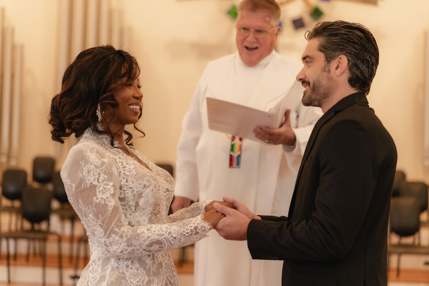 Couple exchanging vows at wedding ceremony with officiant in white robe reading from paper in church.