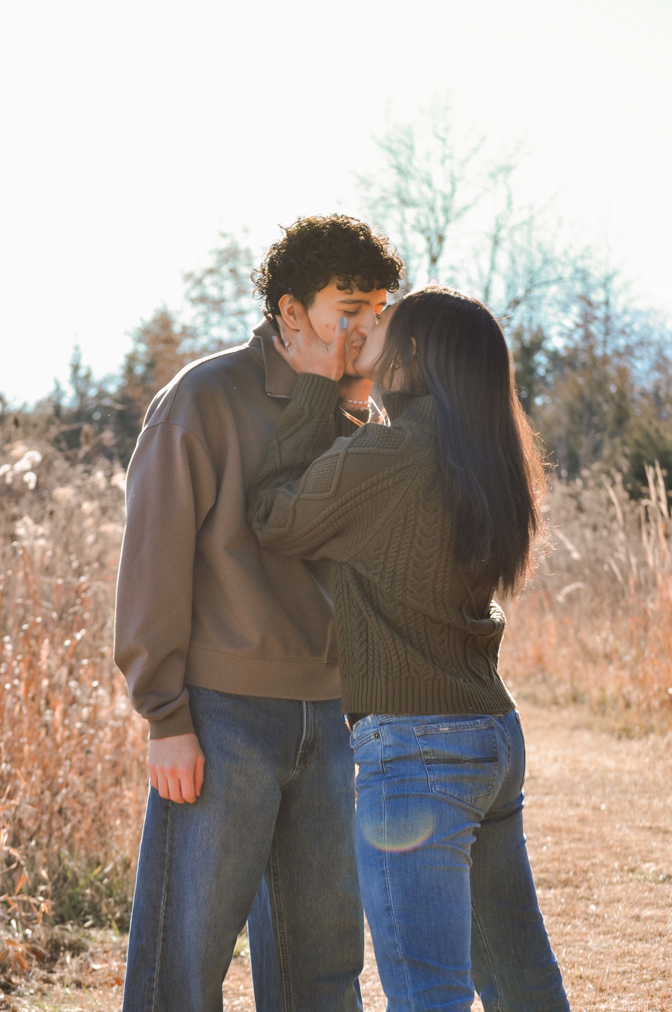 A young couple shares a romantic kiss outdoors in a sunlit field, both wearing casual jeans and sweaters.