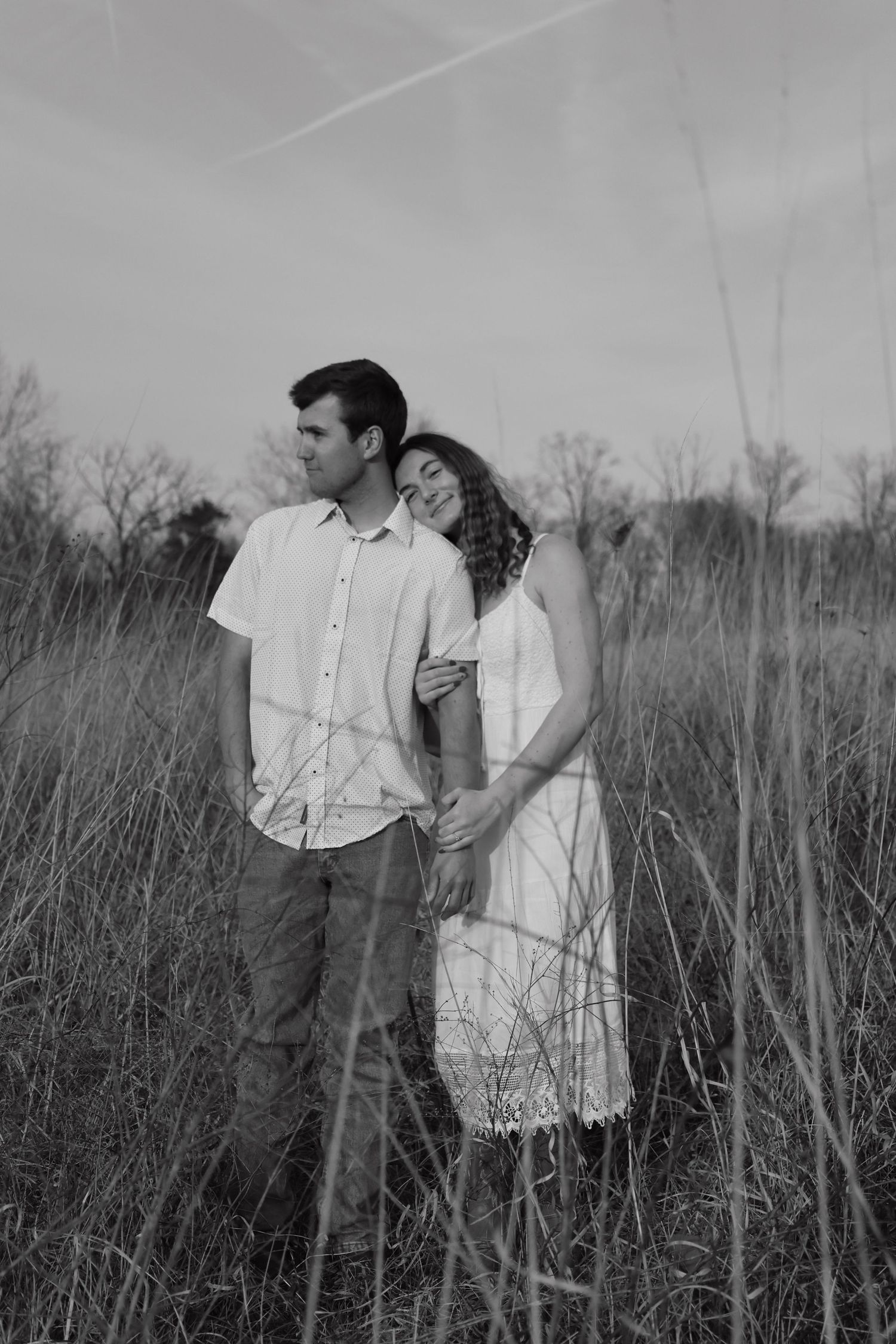 Black and white photo of a couple embracing in a field, woman in white dress leaning on mans shoulder.