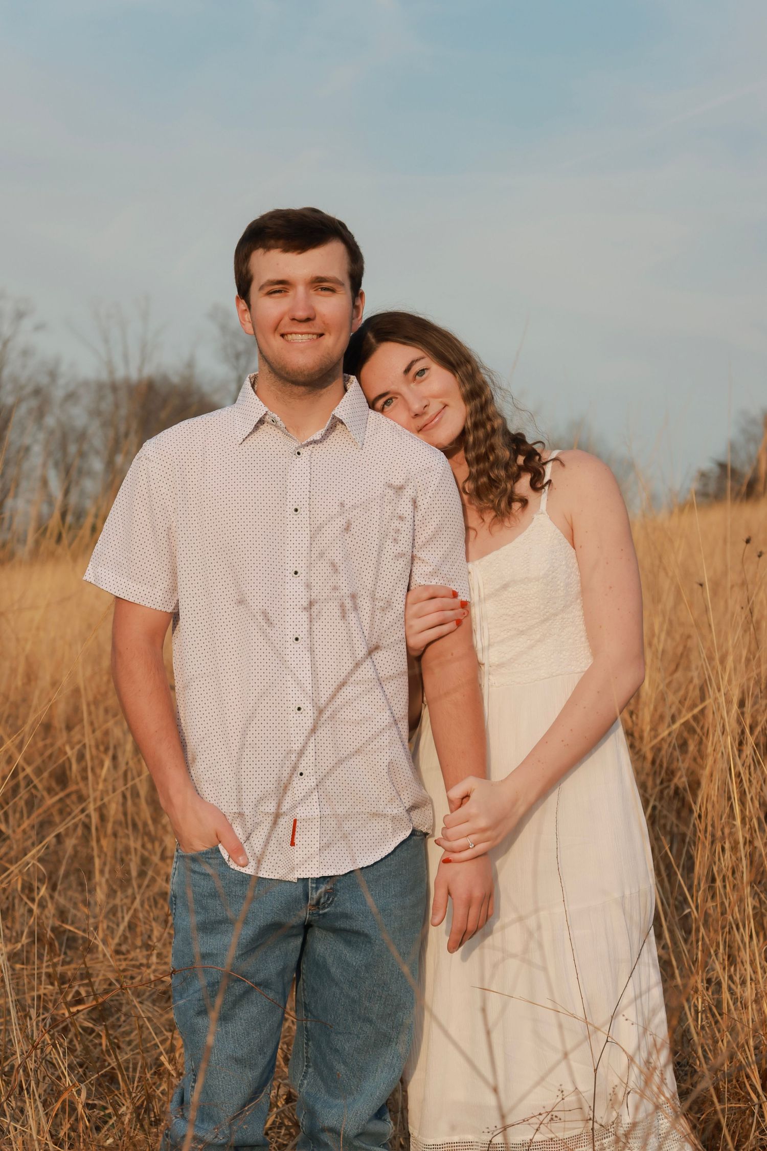 A young couple in white outfits poses in a golden field, the woman leaning on the man's shoulder at sunset.