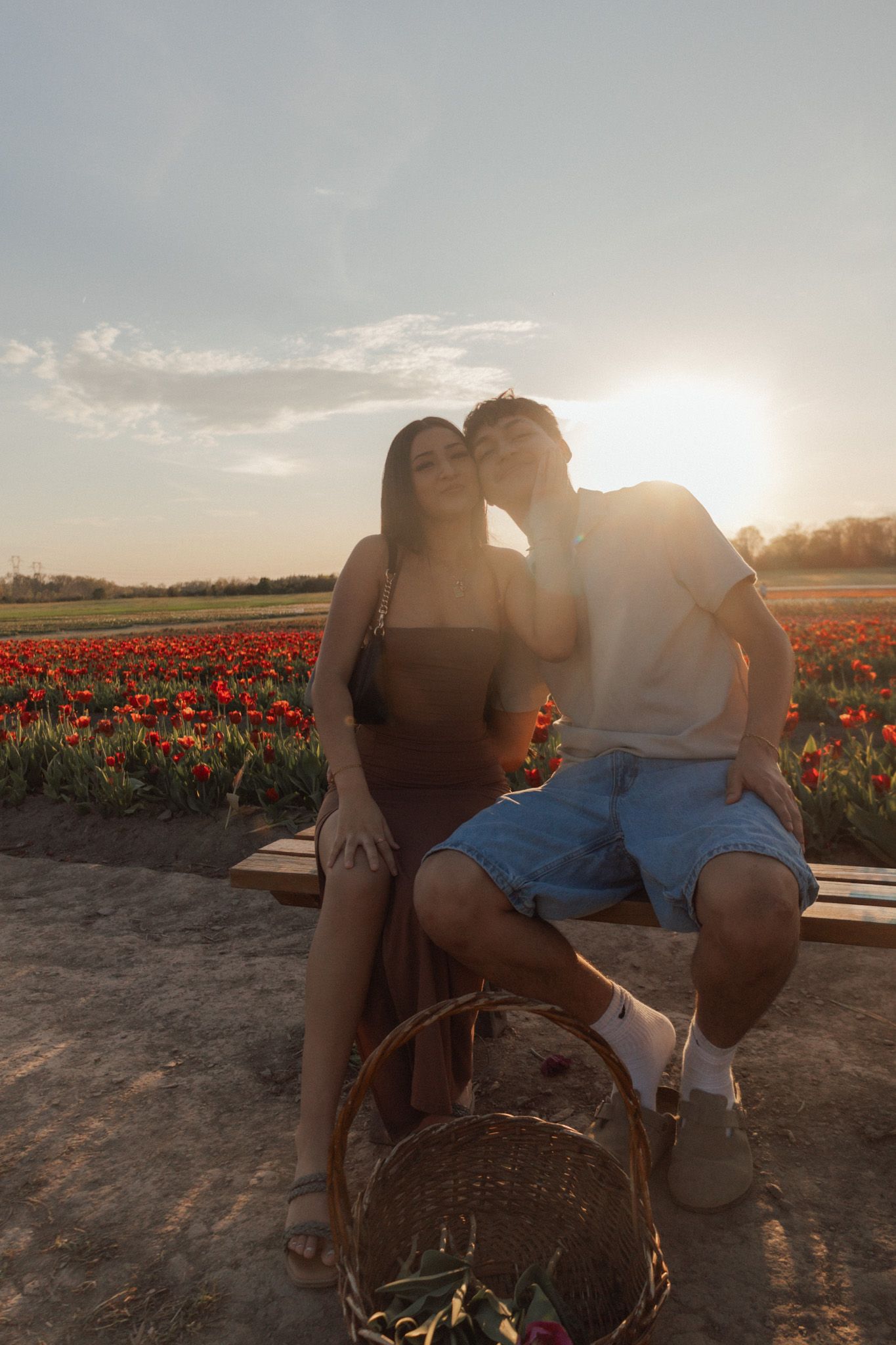 Couple sharing a romantic kiss at sunset in a vibrant red tulip field, with a wicker basket in the foreground.