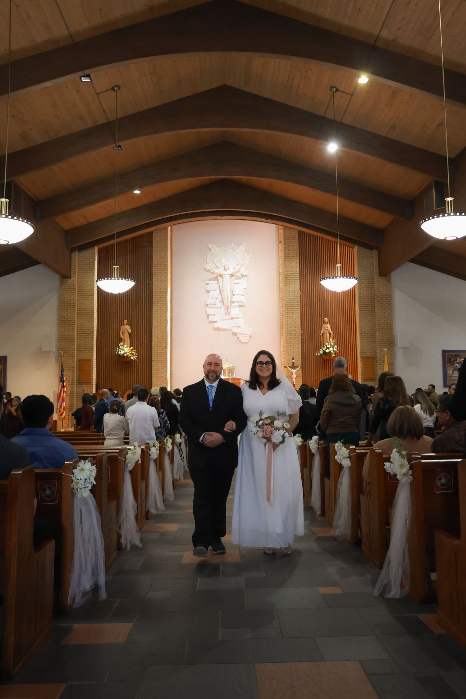 Bride and groom walk down church aisle during wedding ceremony, decorated pews with white bows, wooden cathedral ceiling.