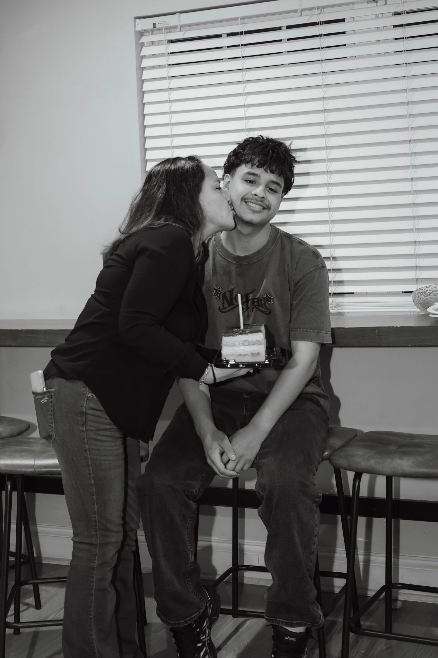 Black and white photo of a young man sitting on a stool smiling as a woman kisses him on the cheek indoors.