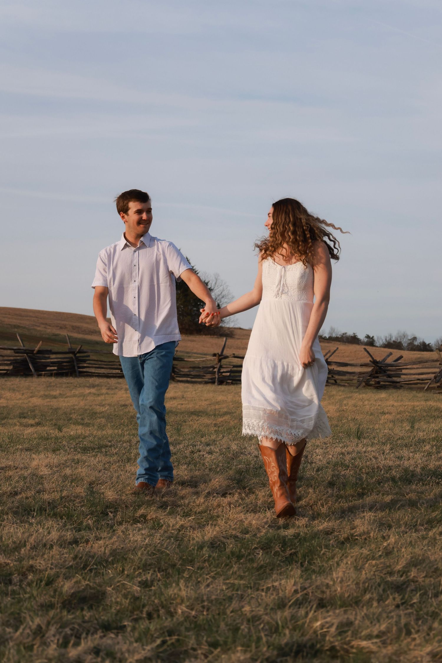 Couple holding hands walking in a golden field with a wooden fence backdrop under a blue sky.