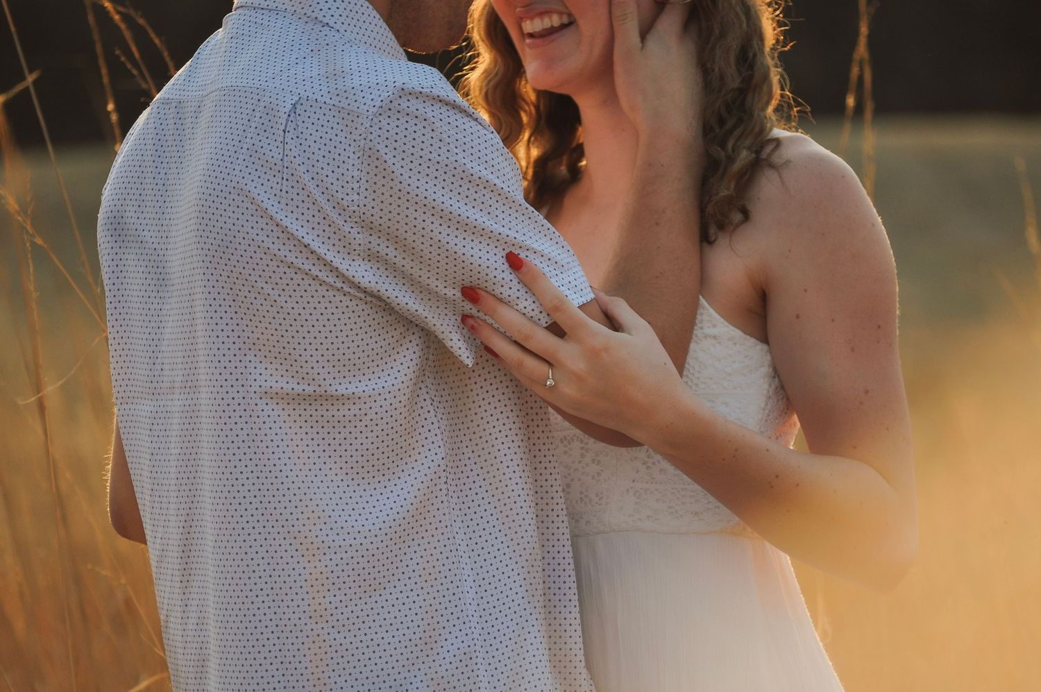Bride smiling and embracing groom in golden field at sunset, wearing white dress with engagement ring visible.