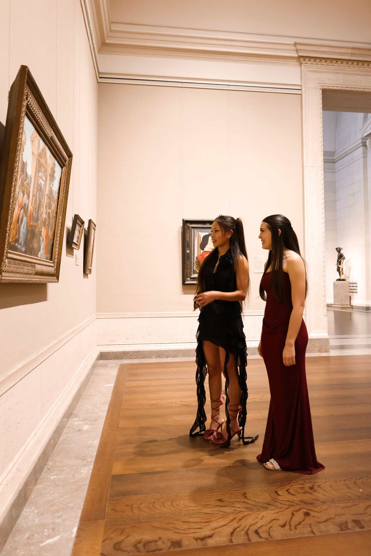 Two elegant women in formal gowns viewing classic paintings in a museum gallery with hardwood floors.
