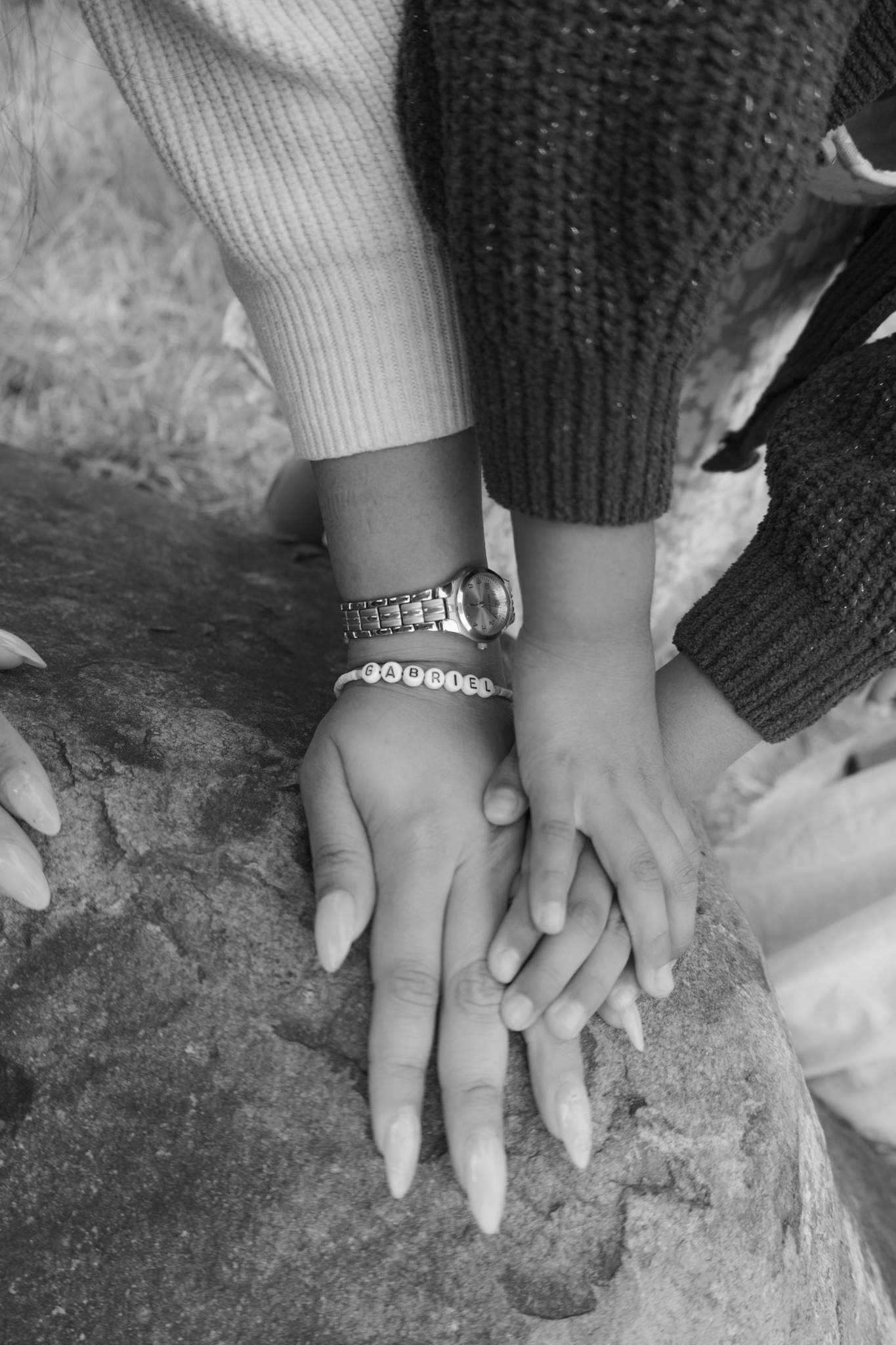 Black and white photo of multiple hands stacked on a rock, one wearing a bracelet lettering gabriel with long nails.