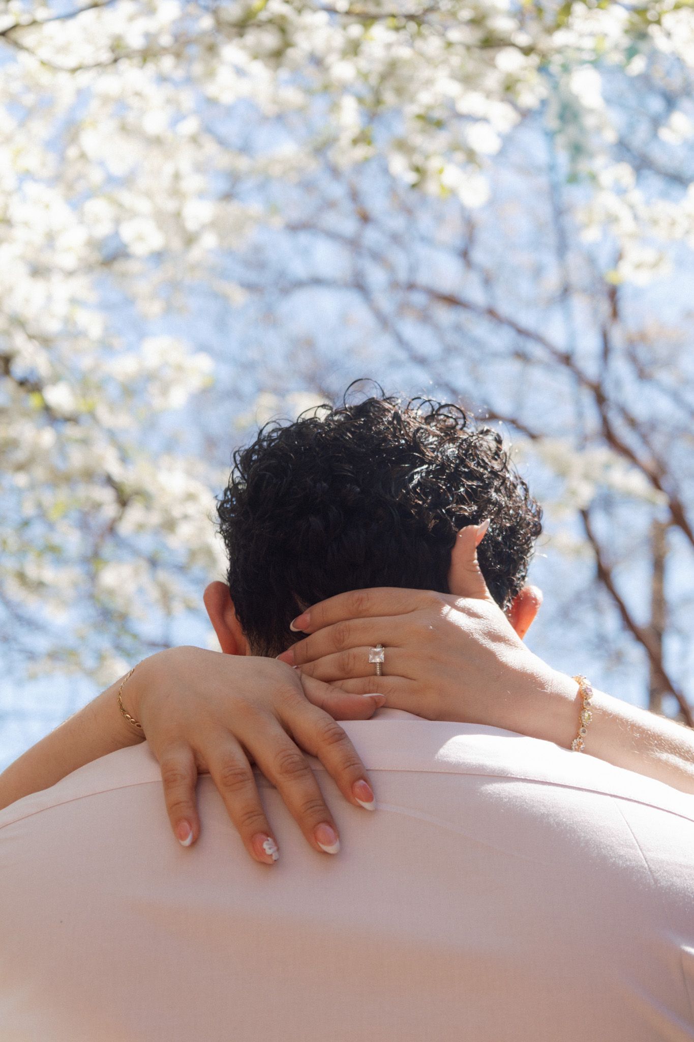 A woman with an engagement ring embraces a man with curly hair under blooming white spring trees.