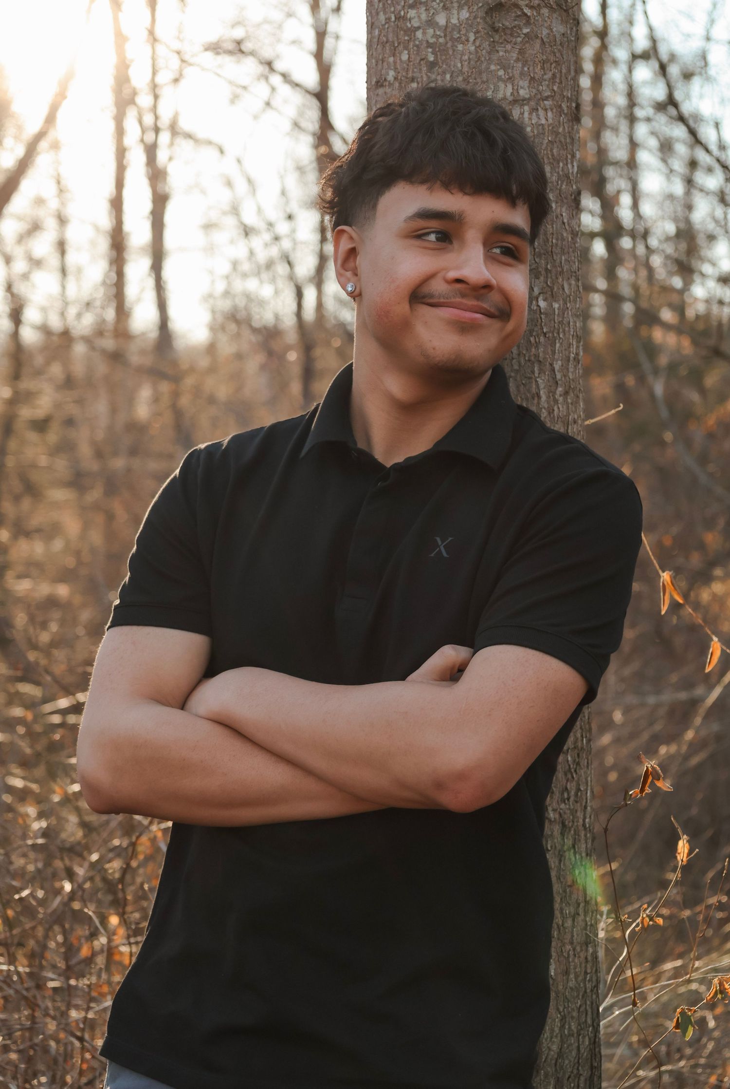 Young man in black polo shirt smiling with arms crossed, leaning against a tree in an autumn forest setting.