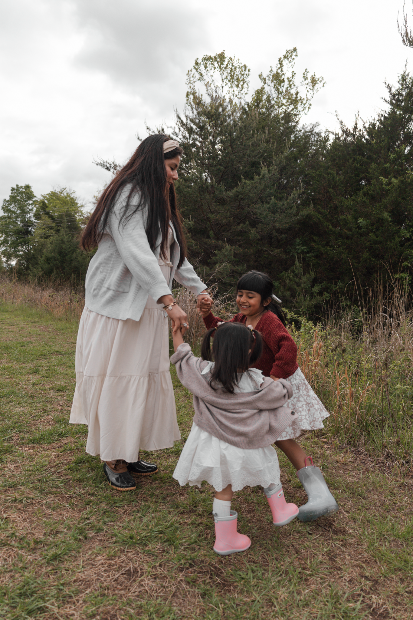 A woman and two young girls hold hands and play together in a grassy field with trees in the background.