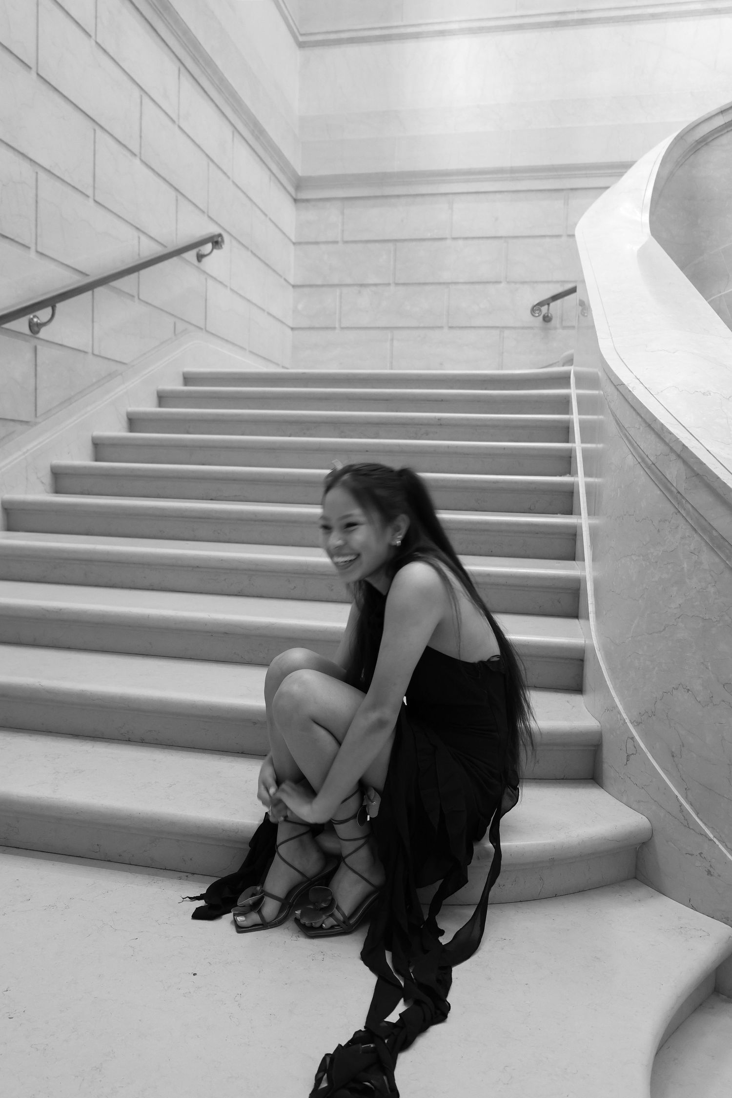 A smiling young woman in a black dress sits gracefully on marble stairs in a black and white photo.
