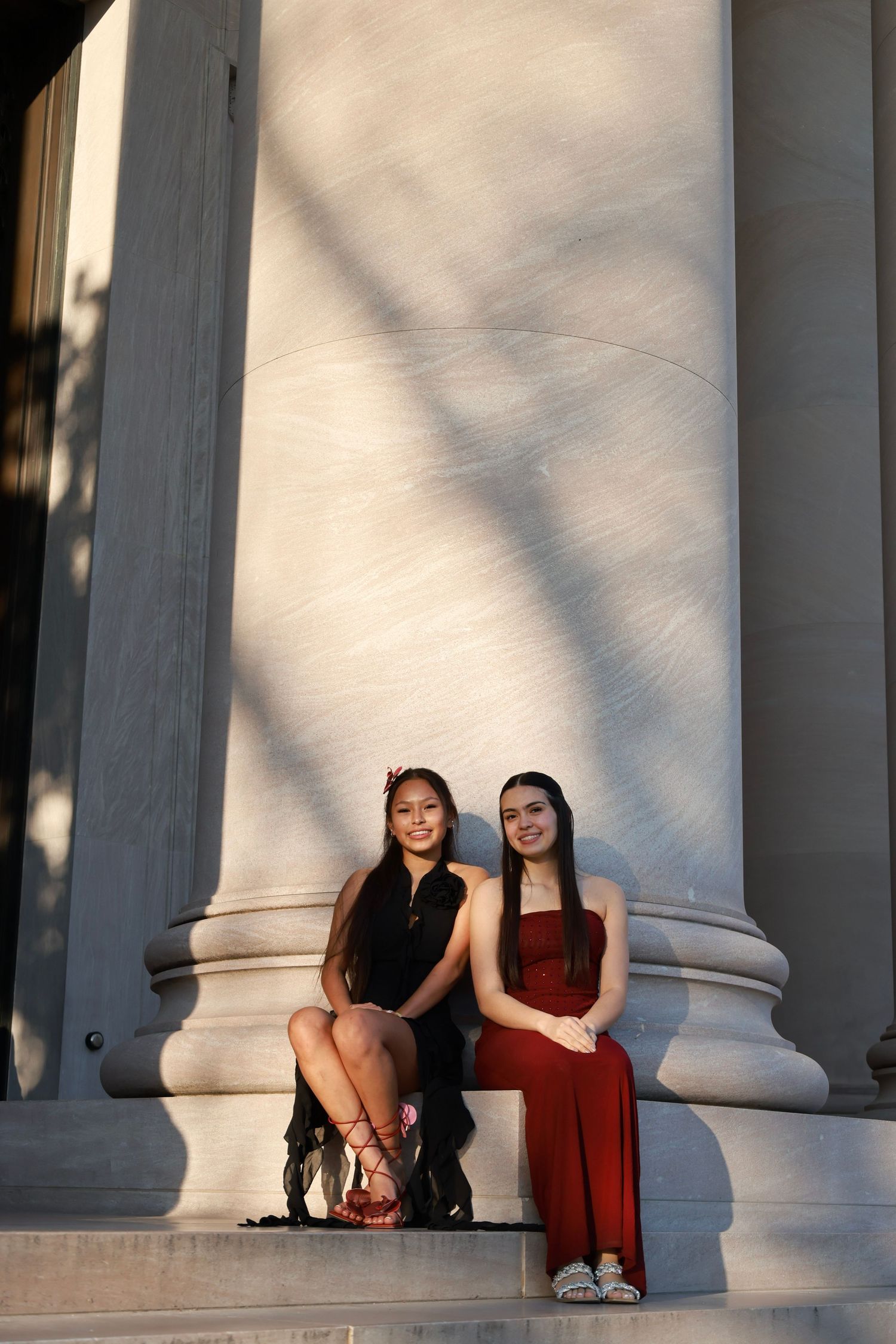 Two young women in elegant red and black formal dresses pose on marble steps between large stone columns.
