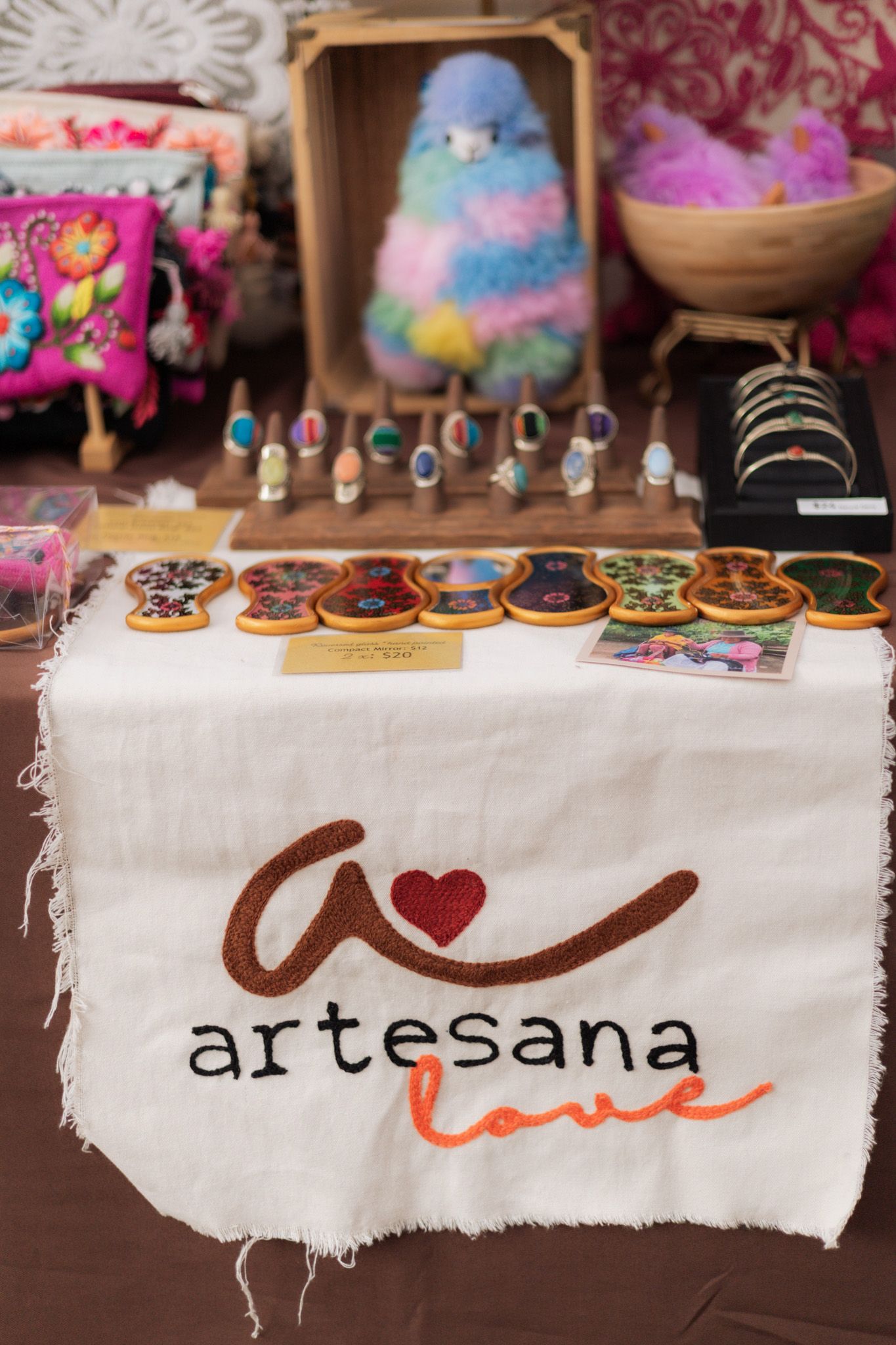 Artesana Love craft market display with colorful alpaca figurine, handmade rings, and decorative wooden ornaments on white cloth.