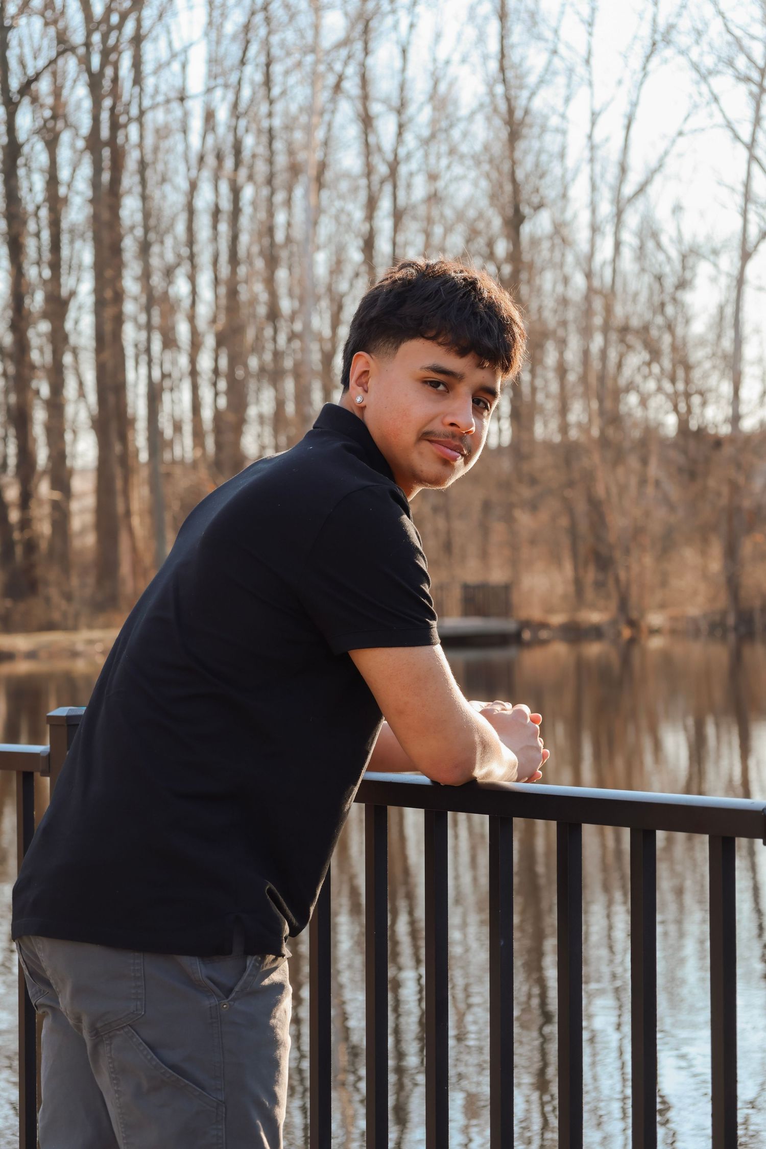 Young man in black polo leans on railing by a serene pond surrounded by bare winter trees on a sunny day.