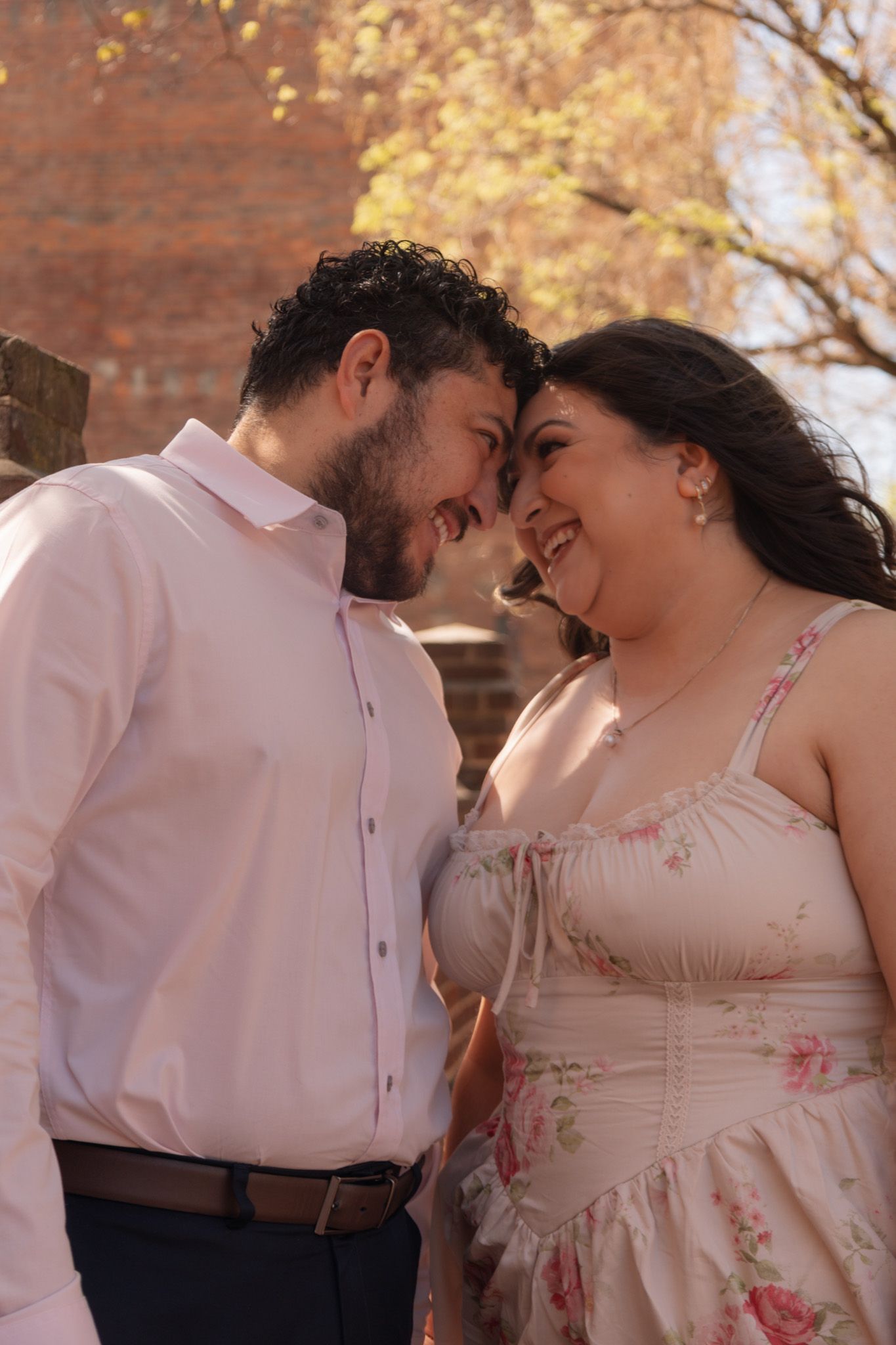 A couple smiles and touches foreheads tenderly outdoors, man in white shirt, woman in floral dress, brick wall behind them.