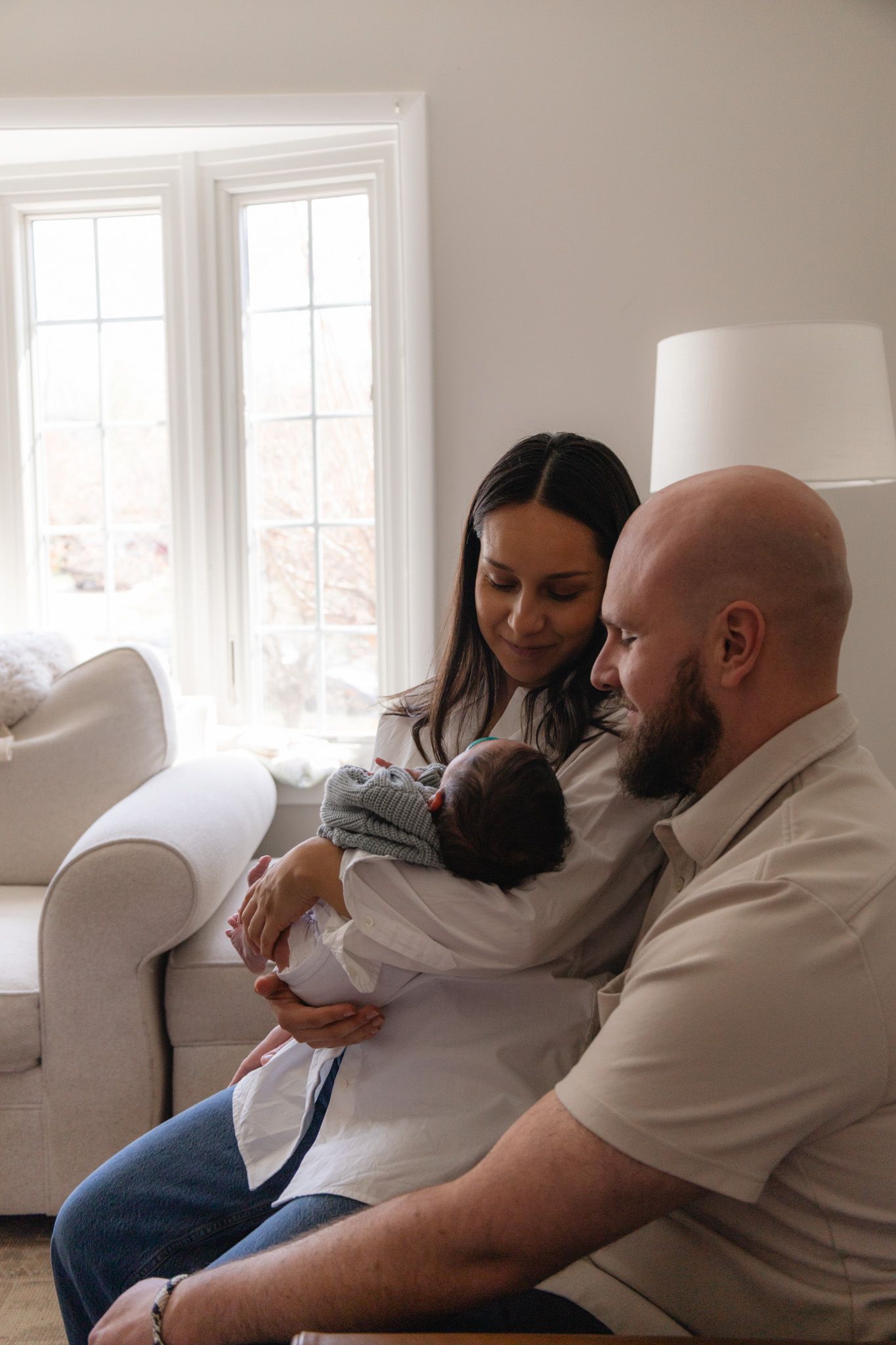 A couple sits together in a bright living room, lovingly gazing at their newborn baby wrapped in a cozy blanket.