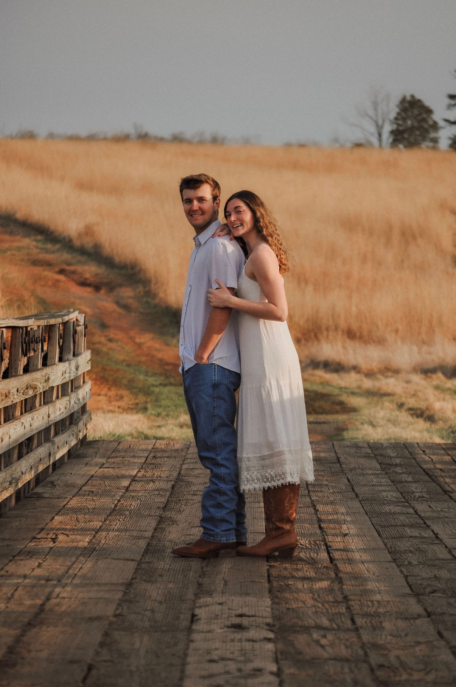 Couple embracing on a wooden bridge surrounded by golden fields during a romantic outdoor engagement photo session.