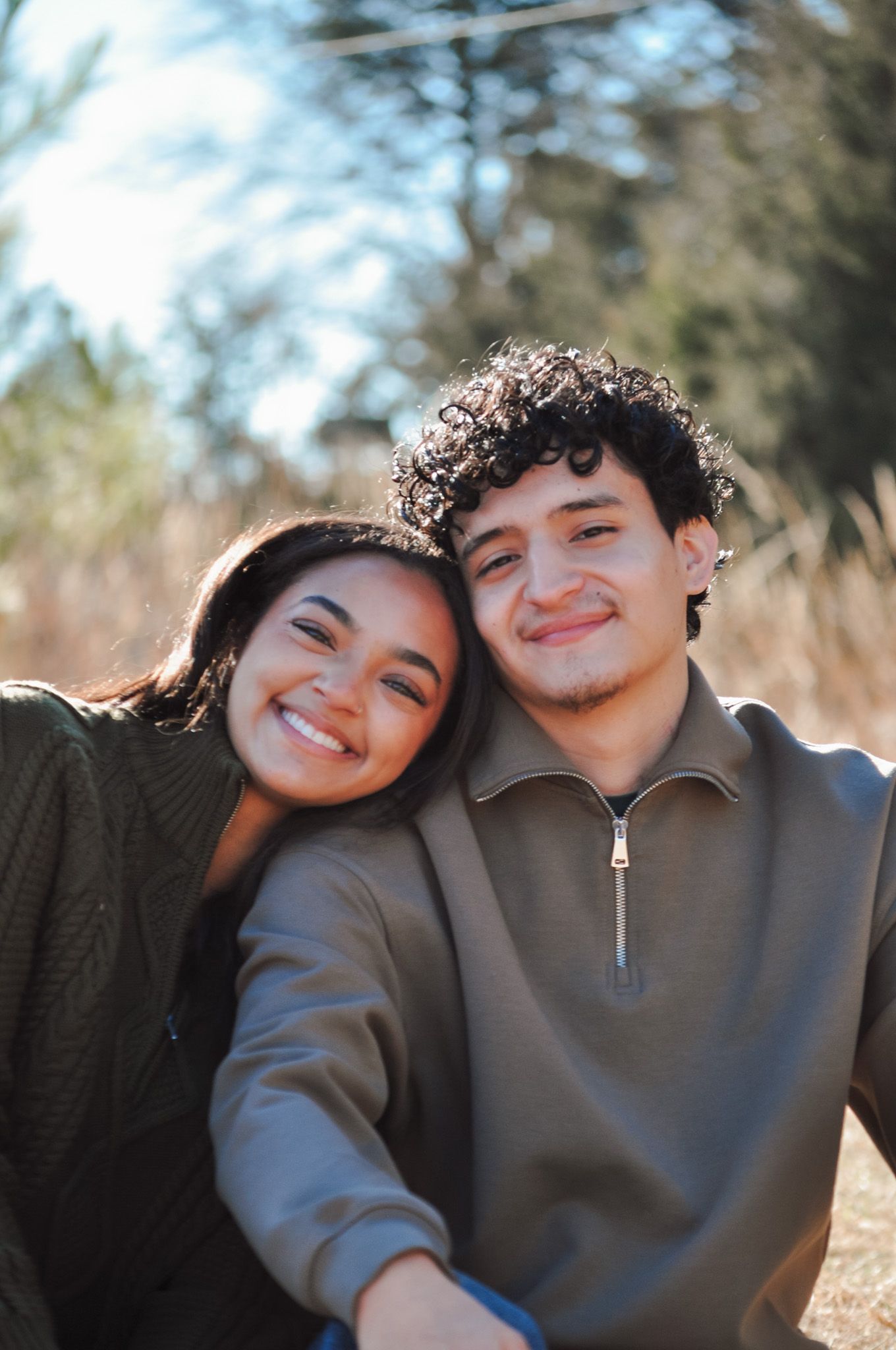 A young couple smiling and posing together outdoors in a sunny natural setting, wearing cozy earth-tone sweaters.
