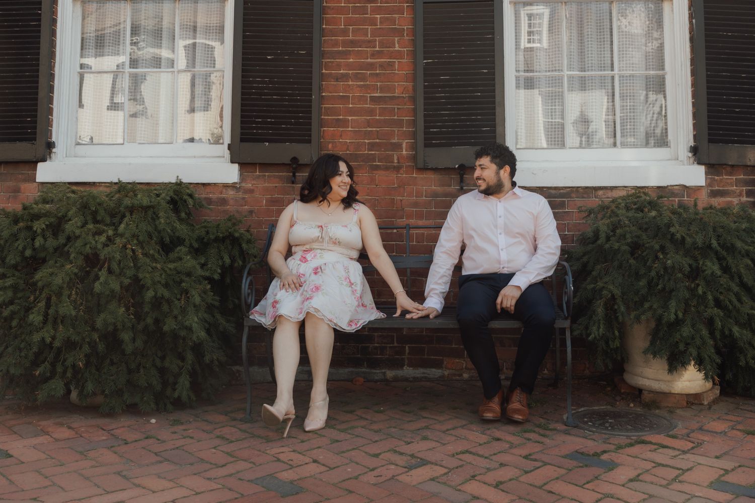 Couple holding hands on a bench outside a brick building with black shutters during an engagement photo session.