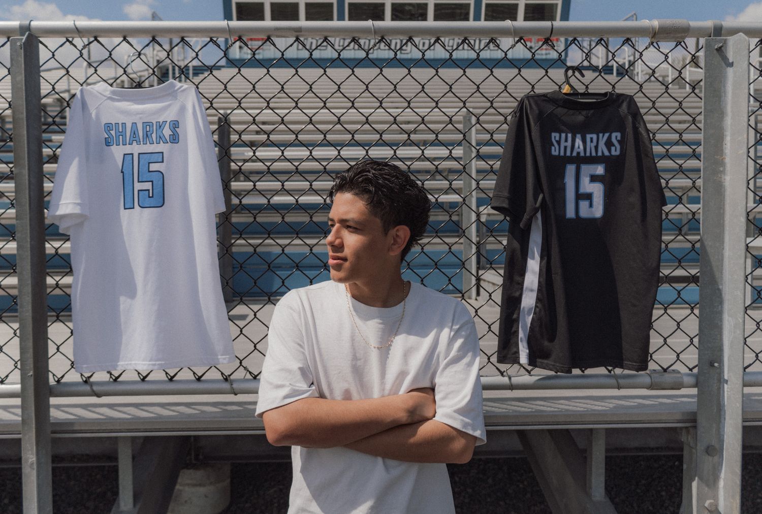 Young athlete poses with arms crossed in front of two Sharks #15 basketball jerseys hung on a chain-link fence.
