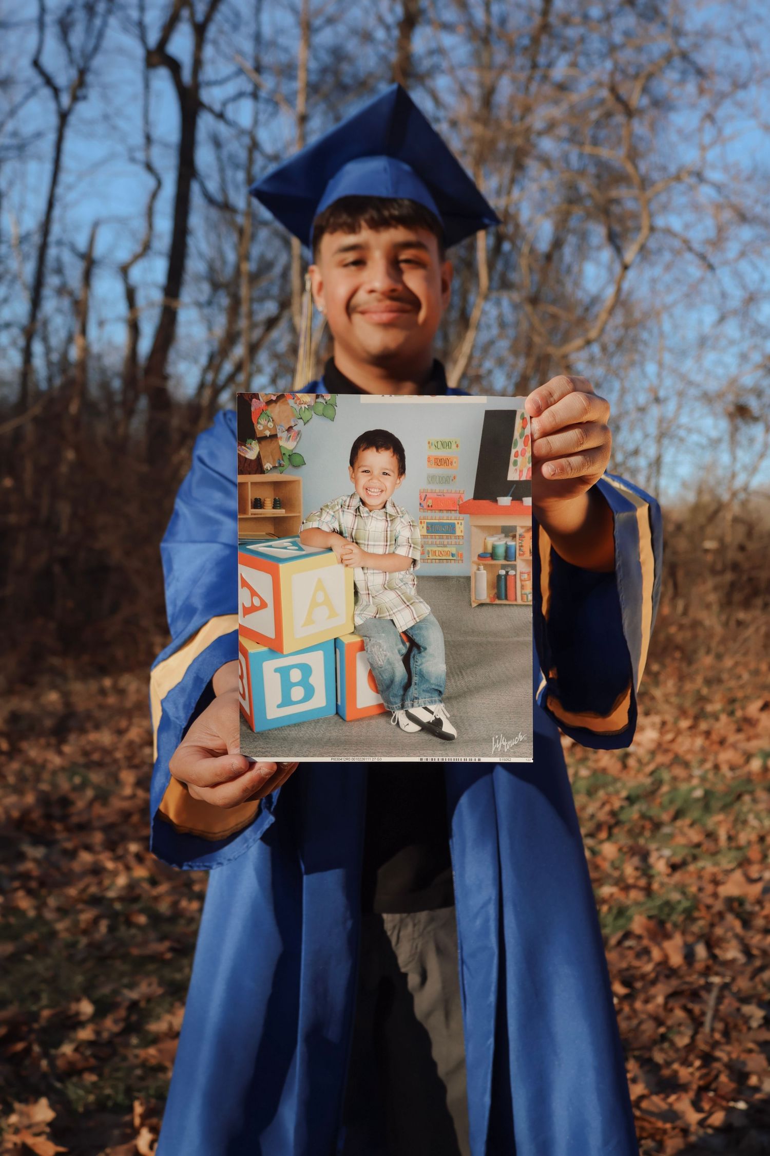 A smiling graduate in blue cap and gown holds a childhood kindergarten photo outdoors among autumn trees.