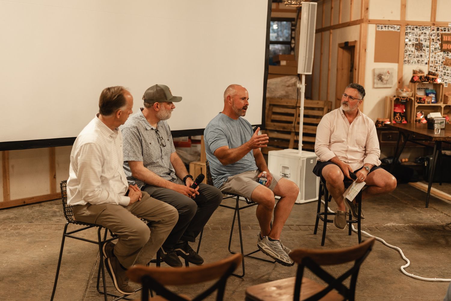 Four men seated on stools in casual panel discussion inside rustic venue with white projection screen backdrop.