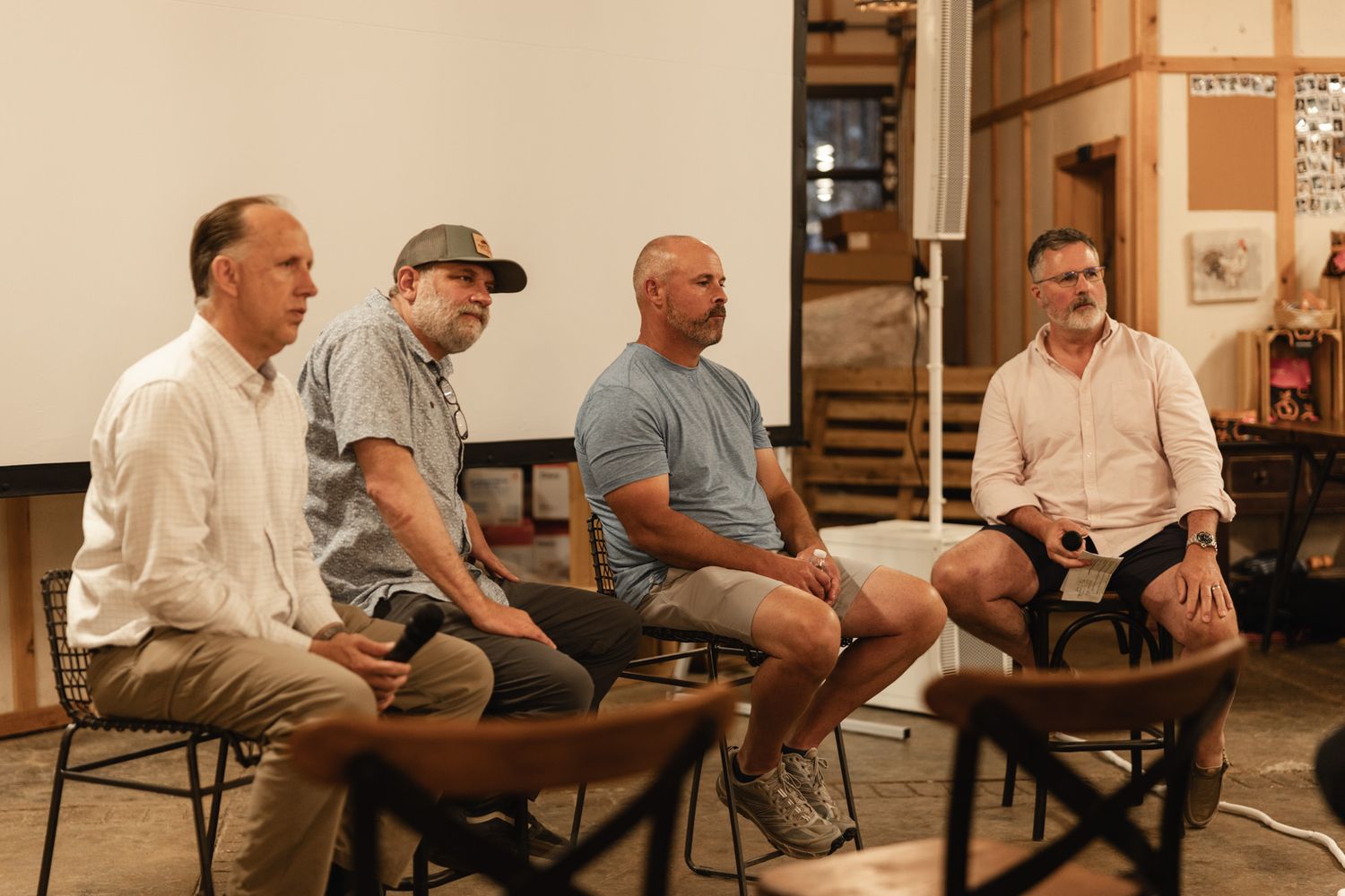 Four men seated in chairs during an indoor panel discussion in a rustic venue setting.